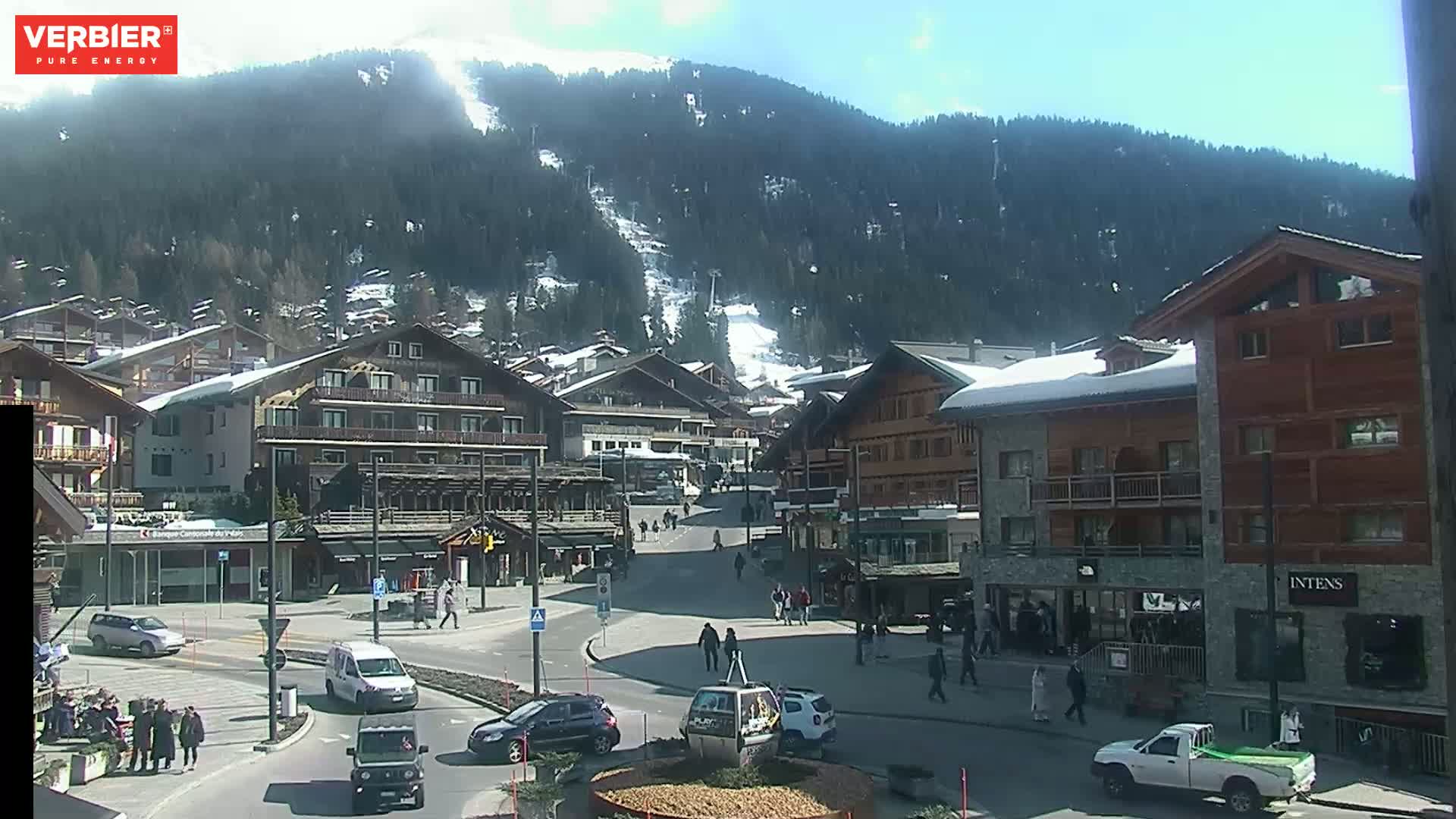 A sunny day in a snow-dusted mountain village, showing a town square with several buildings, cars, and pedestrians, with a ski slope visible on a mountain in the background.