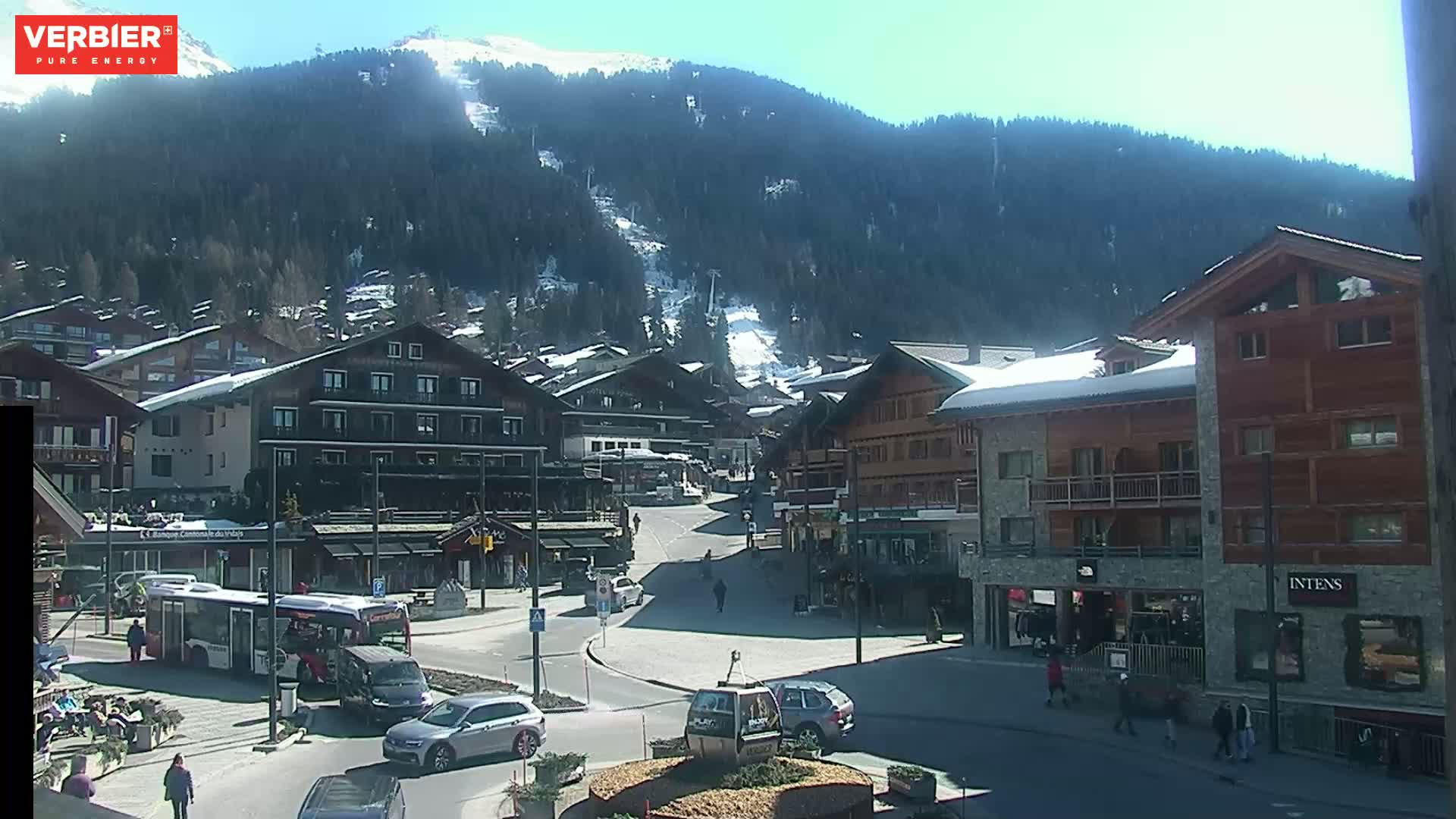 A sunny day in a snow-covered mountain village with several buildings, a road, cars, and pedestrians.
