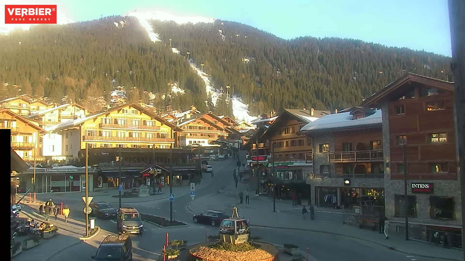 A sunny, snow-dusted mountainside overlooks a village square with several multi-story buildings, a few cars, and pedestrians.