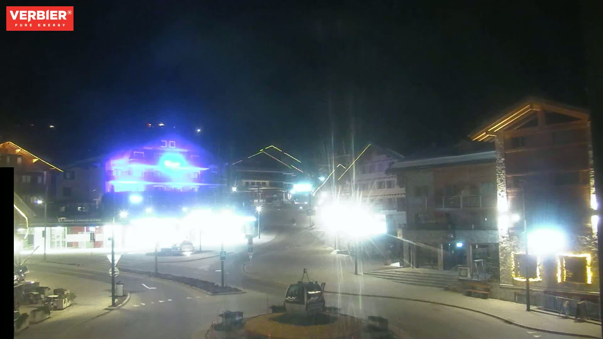 A nighttime view of a town square with brightly lit buildings and streetlights, under a clear, dark sky.