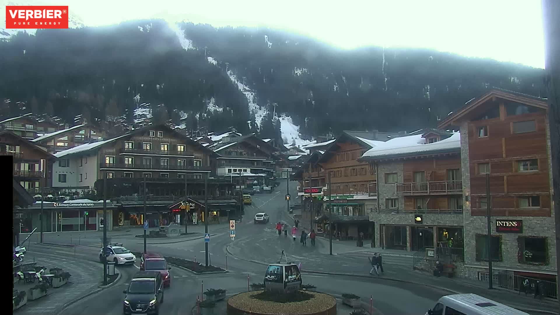 A snow-dusted village square with cars and pedestrians, situated at the base of a snow-covered mountain under an overcast sky.