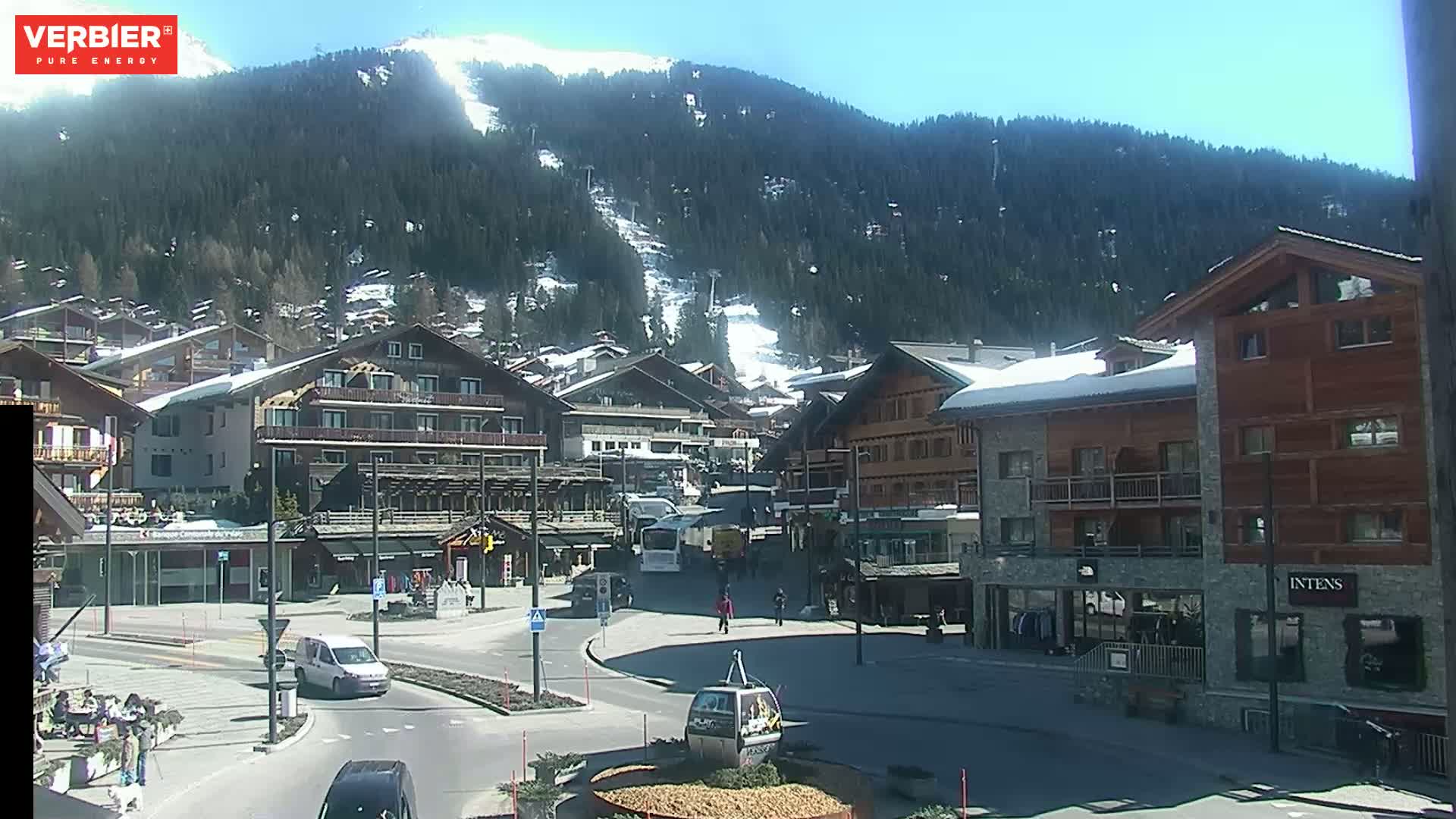 A sunny day in a village nestled at the base of a snow-covered mountain, with several buildings and a few cars visible in a town square.