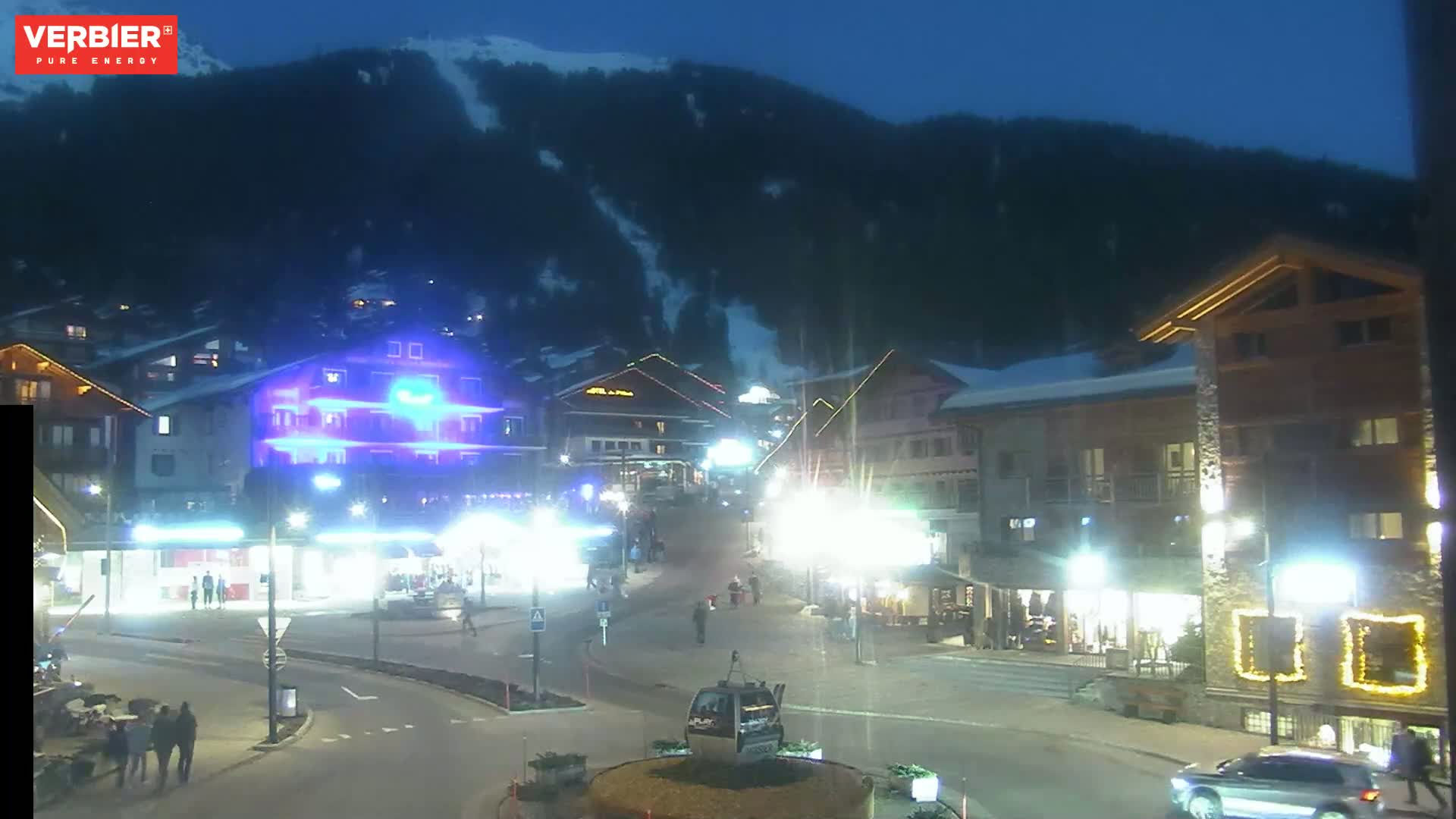 A nighttime view of a snow-covered mountain overlooking a brightly lit town square with several buildings and people walking around under a dark, clear sky.