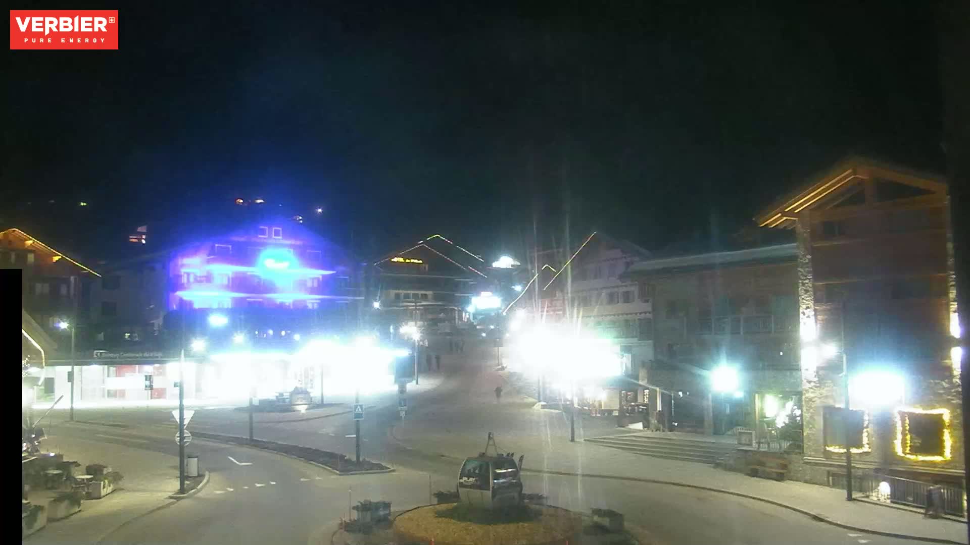 A nighttime, brightly lit town square with several buildings, streetlights, and a cable car in the center.