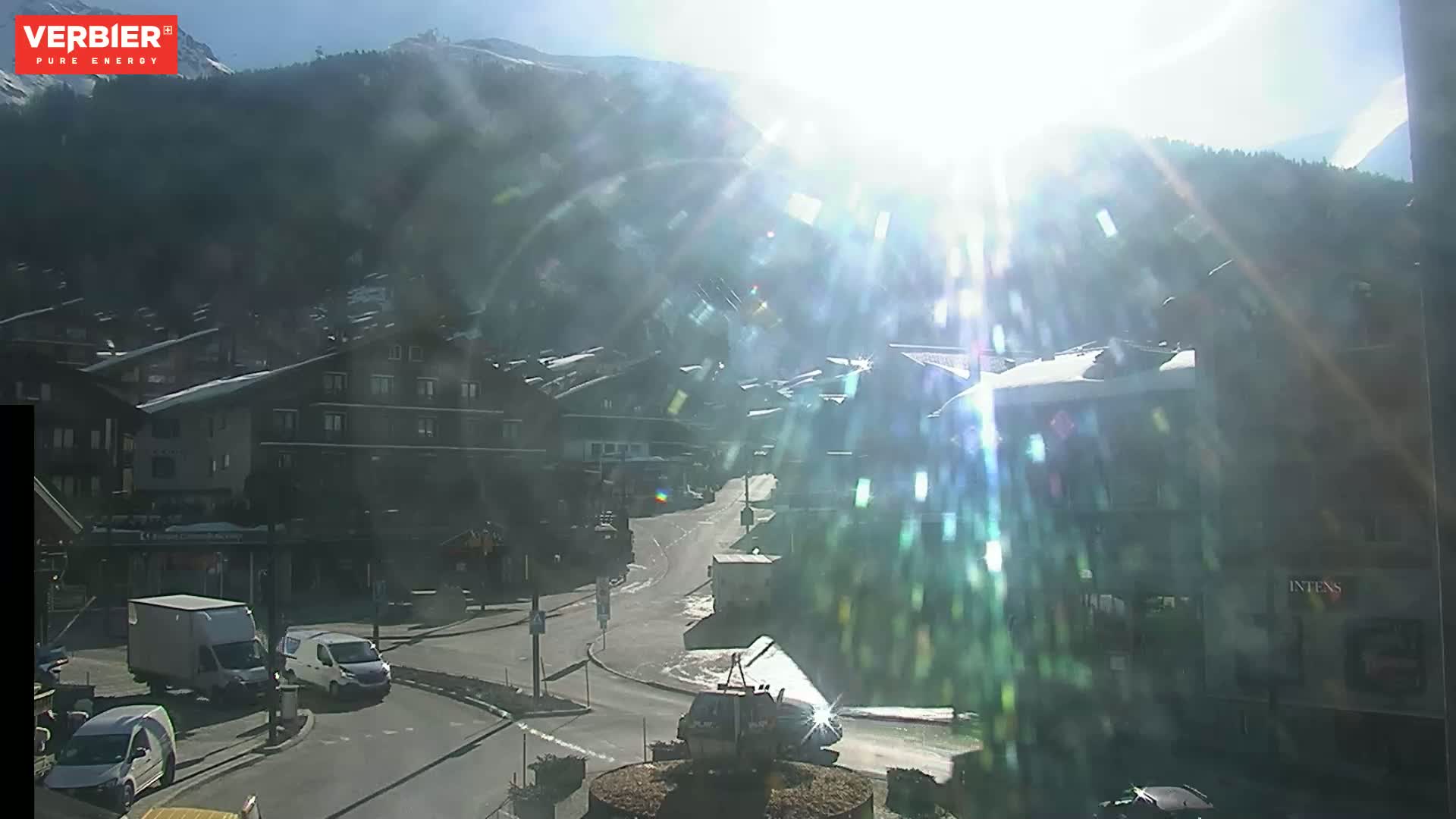A bright, sunny day reveals a village nestled in a valley, with snow-capped mountains in the background and several vehicles parked and driving on a road.