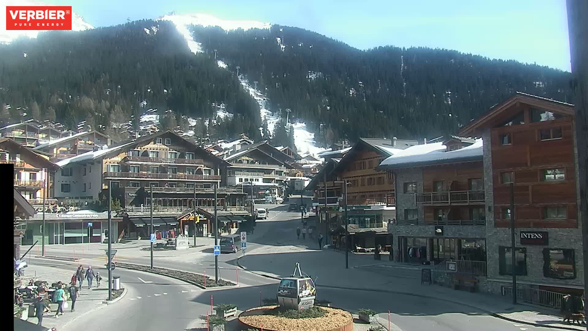 A sunny day reveals a village nestled in a valley, with snow-covered mountains in the background and several people walking on the streets.