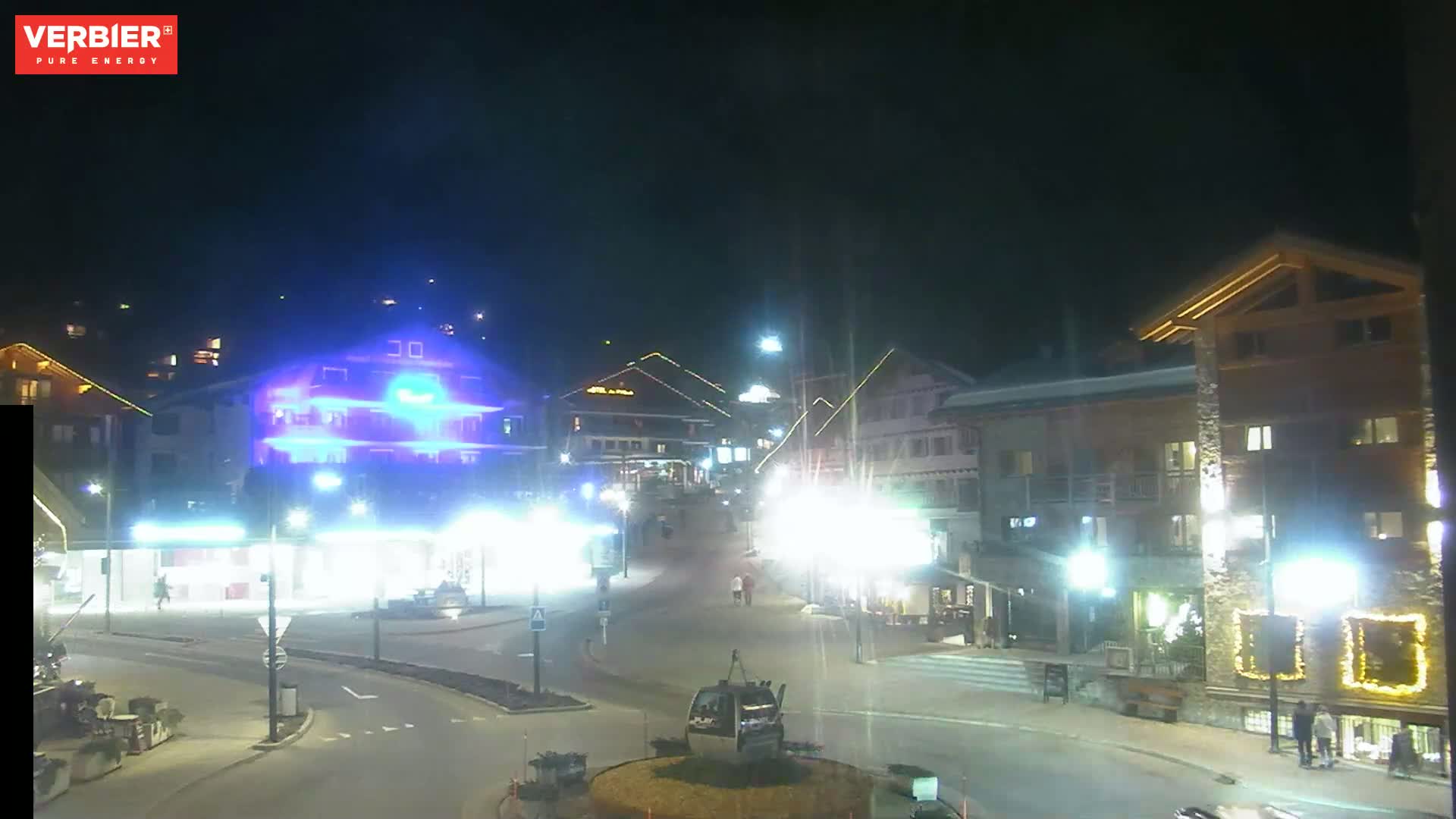 A nighttime view of a town square with brightly lit buildings, streetlights, and a cable car gondola, under clear skies.