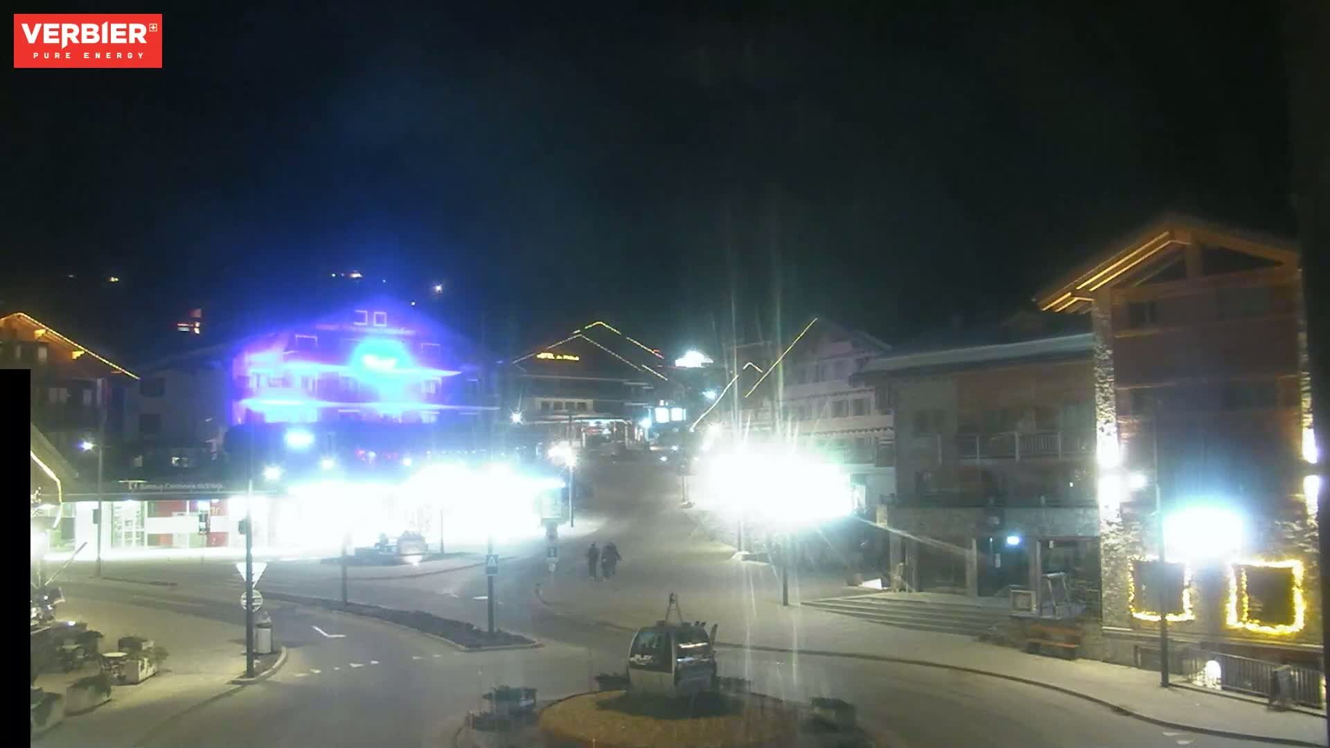 A nighttime view of a town square with several buildings illuminated, a gondola lift in the center, and a few pedestrians.