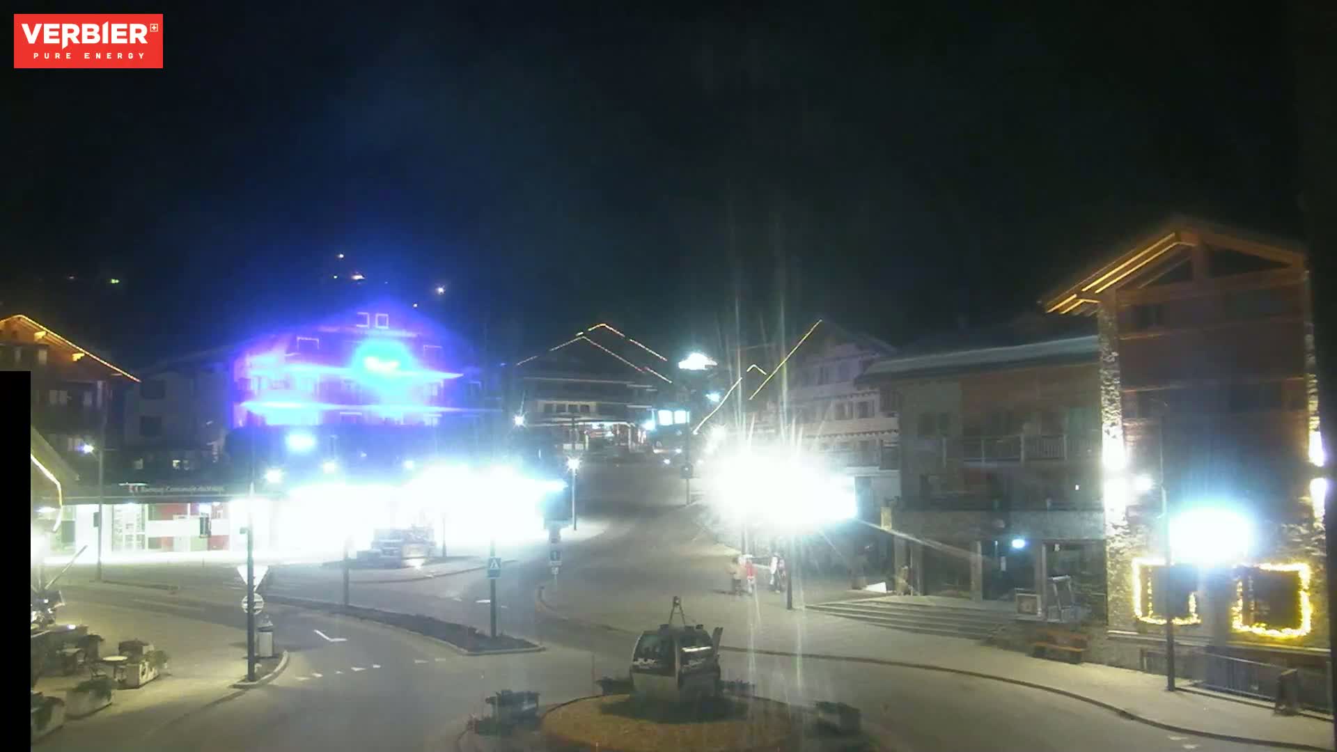A nighttime view of a town square with brightly lit buildings and a few pedestrians, under a clear, dark sky.