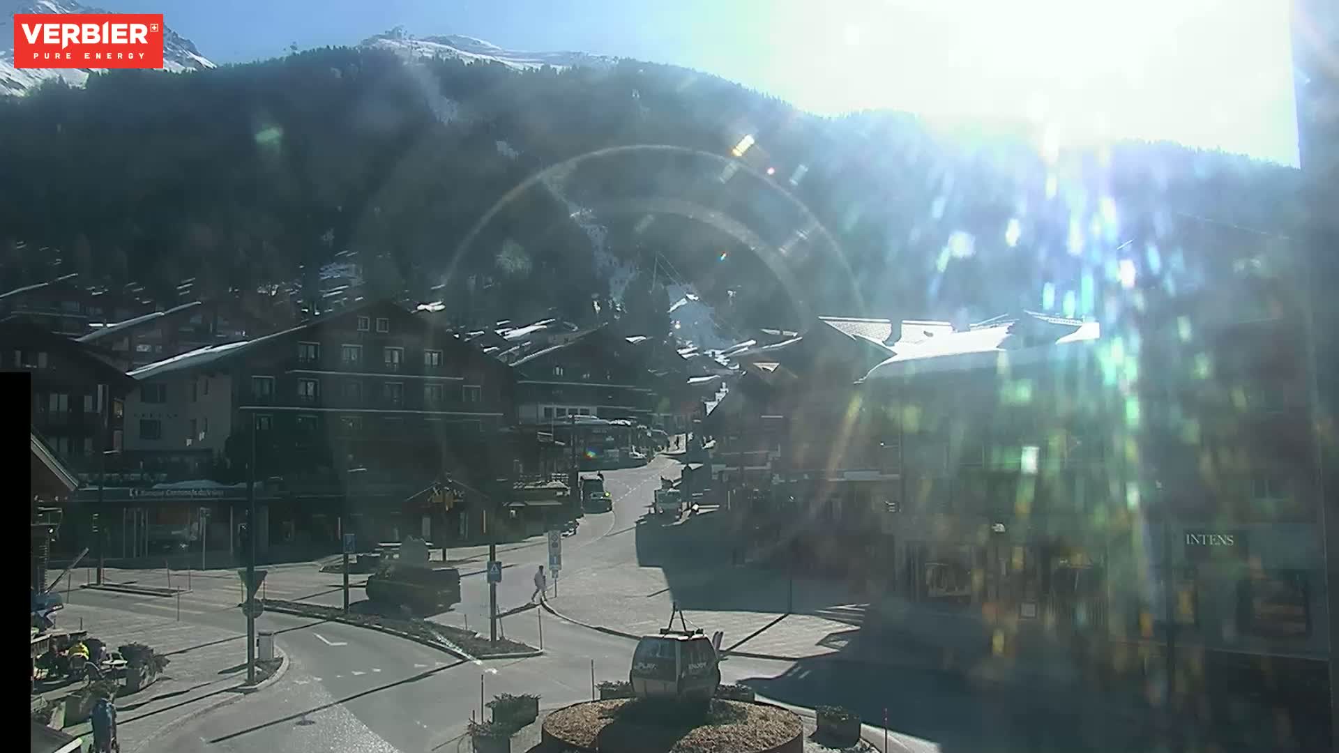 A sunny day in a snow-covered mountain village shows a town square with buildings, vehicles, and a cable car, with bright sunlight creating lens flare.