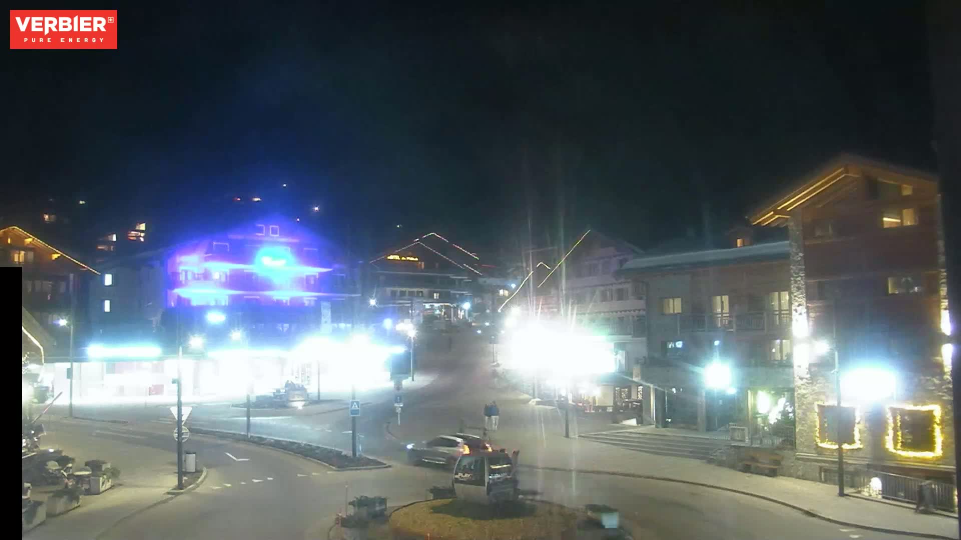 A nighttime view of a town square with brightly lit buildings, a few cars, and a generally clear sky.