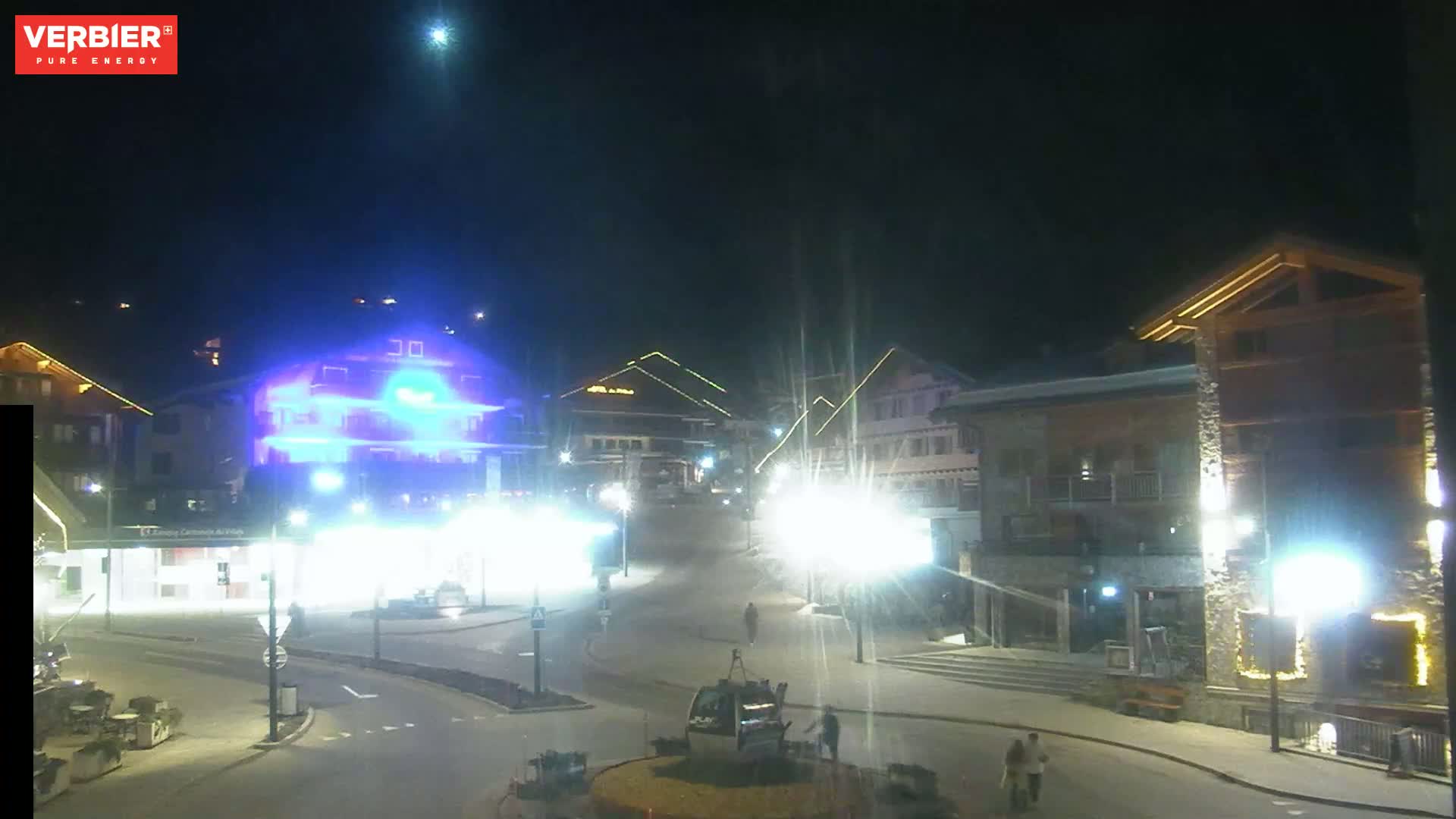 A nighttime view of a town square with brightly lit buildings, streetlights, and a gondola lift, under a clear night sky.