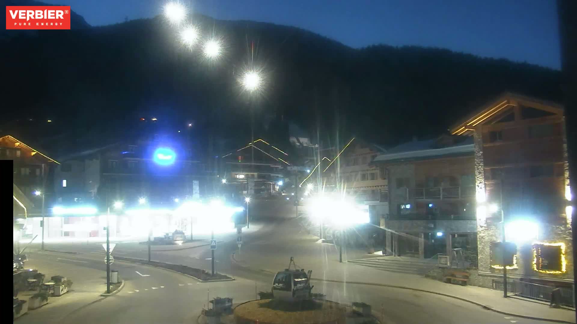 A brightly lit town square at night, nestled against a dark mountainside under a clear, twilight sky.