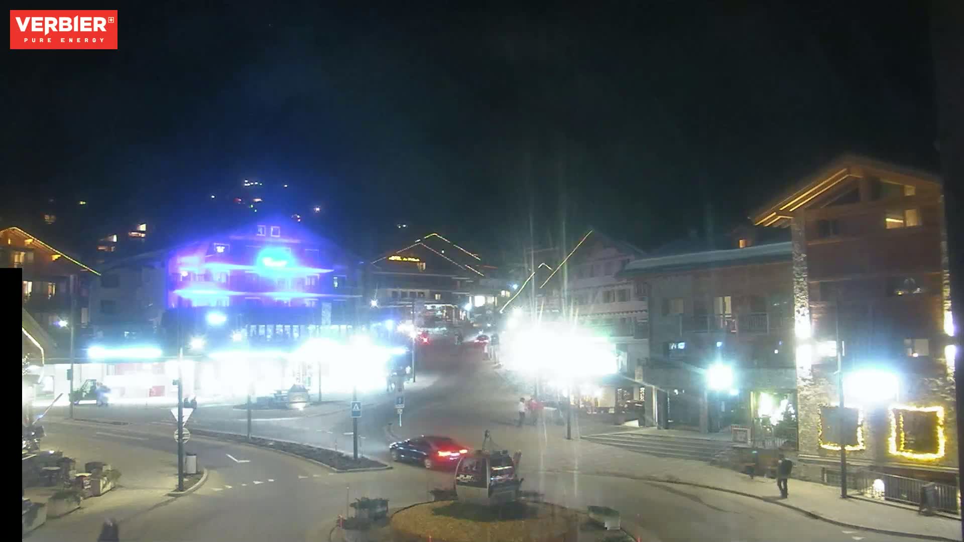 A nighttime view of a town square with several buildings illuminated, a few cars, and pedestrians walking around.
