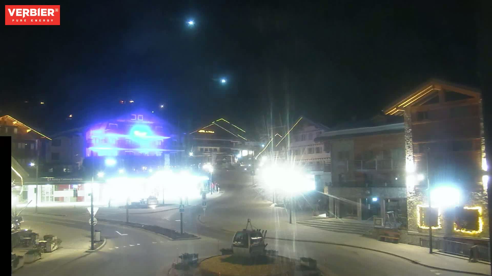 A nighttime view of a town square with brightly lit buildings and a gondola lift, under a clear, dark sky.