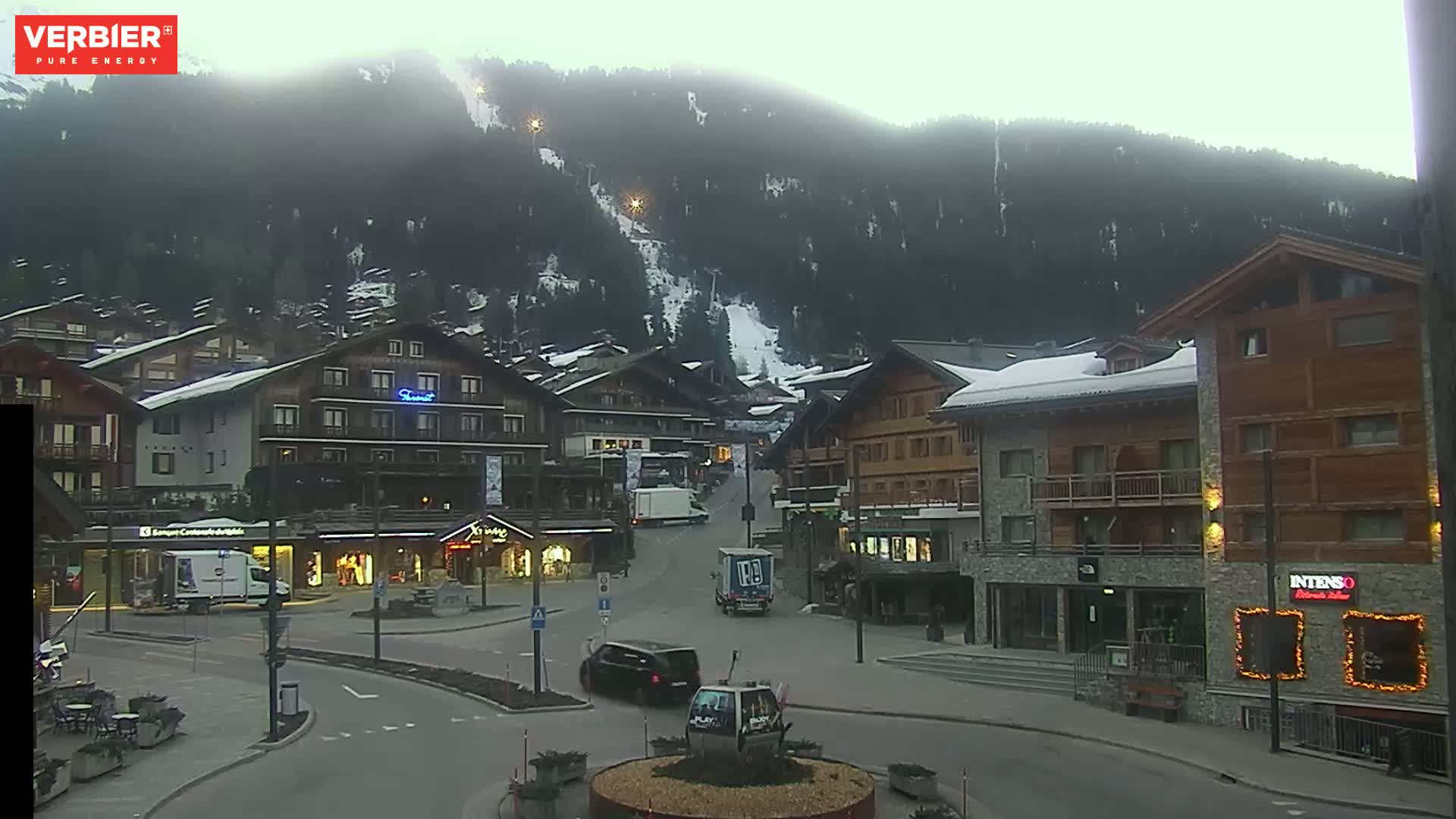 A town square in a snow-dusted mountain village, with overcast skies and several vehicles parked and driving through.