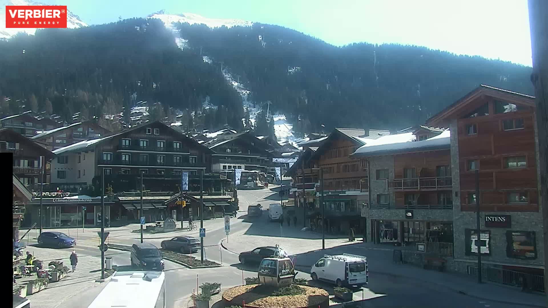 A sunny day in a snow-covered mountain town shows a village square with cars and buildings, and a mountainside with ski runs in the background.