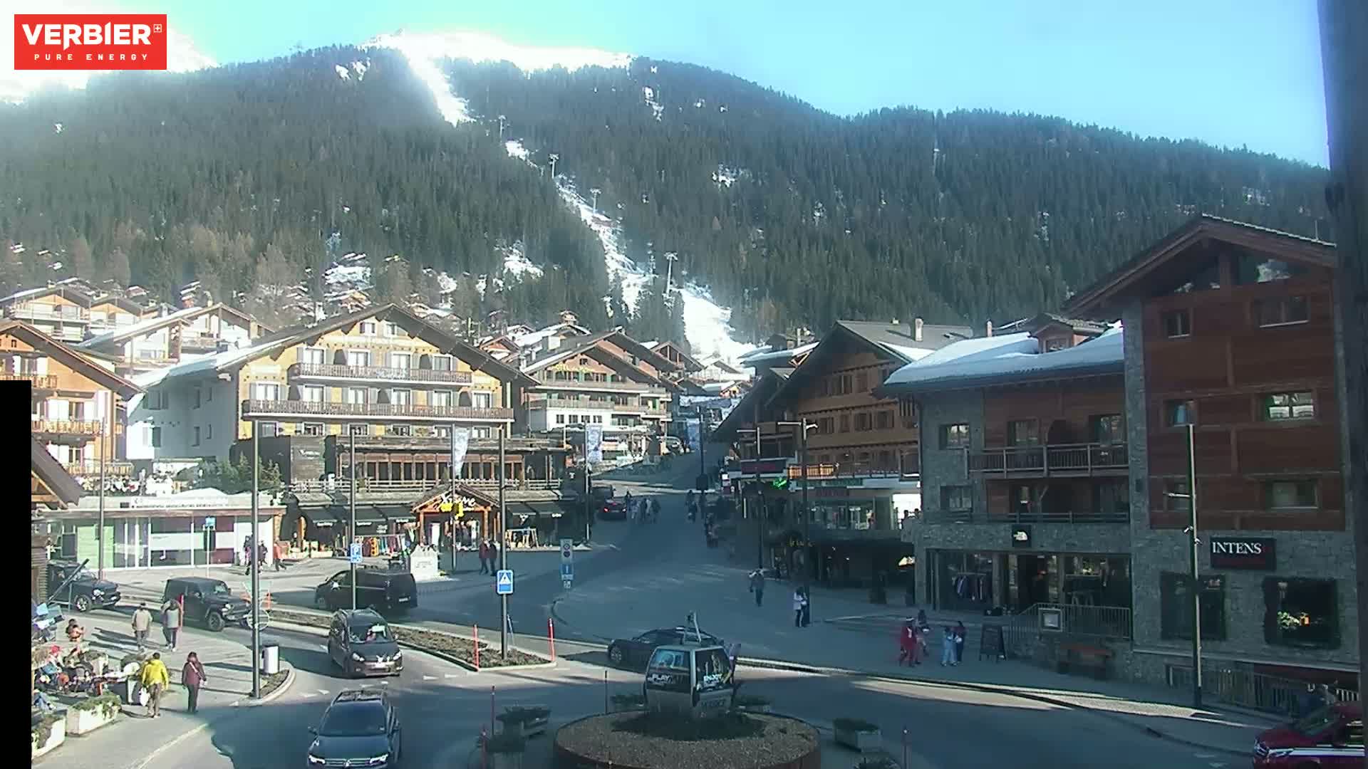 A sunny day in a village nestled in a valley, surrounded by snow-dusted mountains and evergreen trees, with cars parked along a street and people walking around.