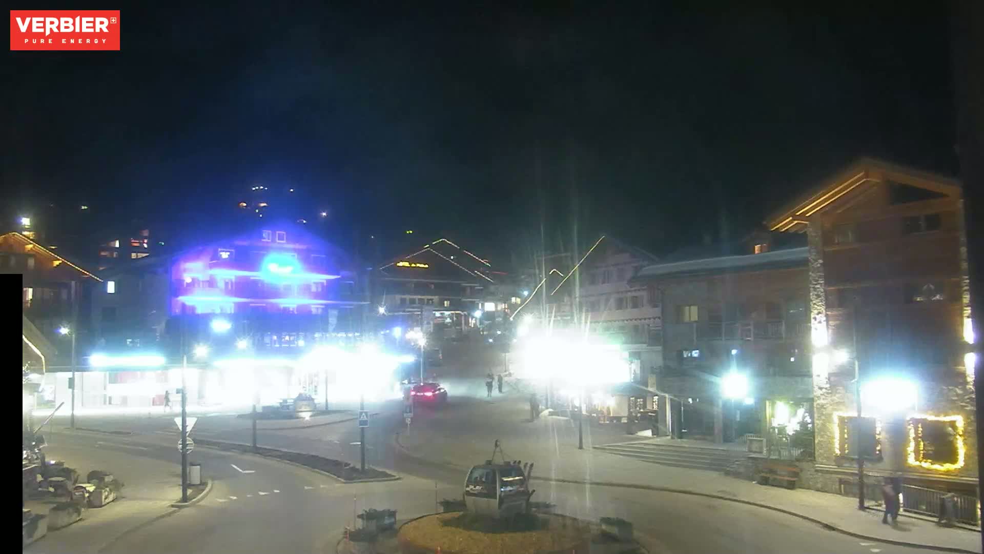 A nighttime view of a town square with brightly lit buildings and a few cars and people visible under a dark, clear sky.