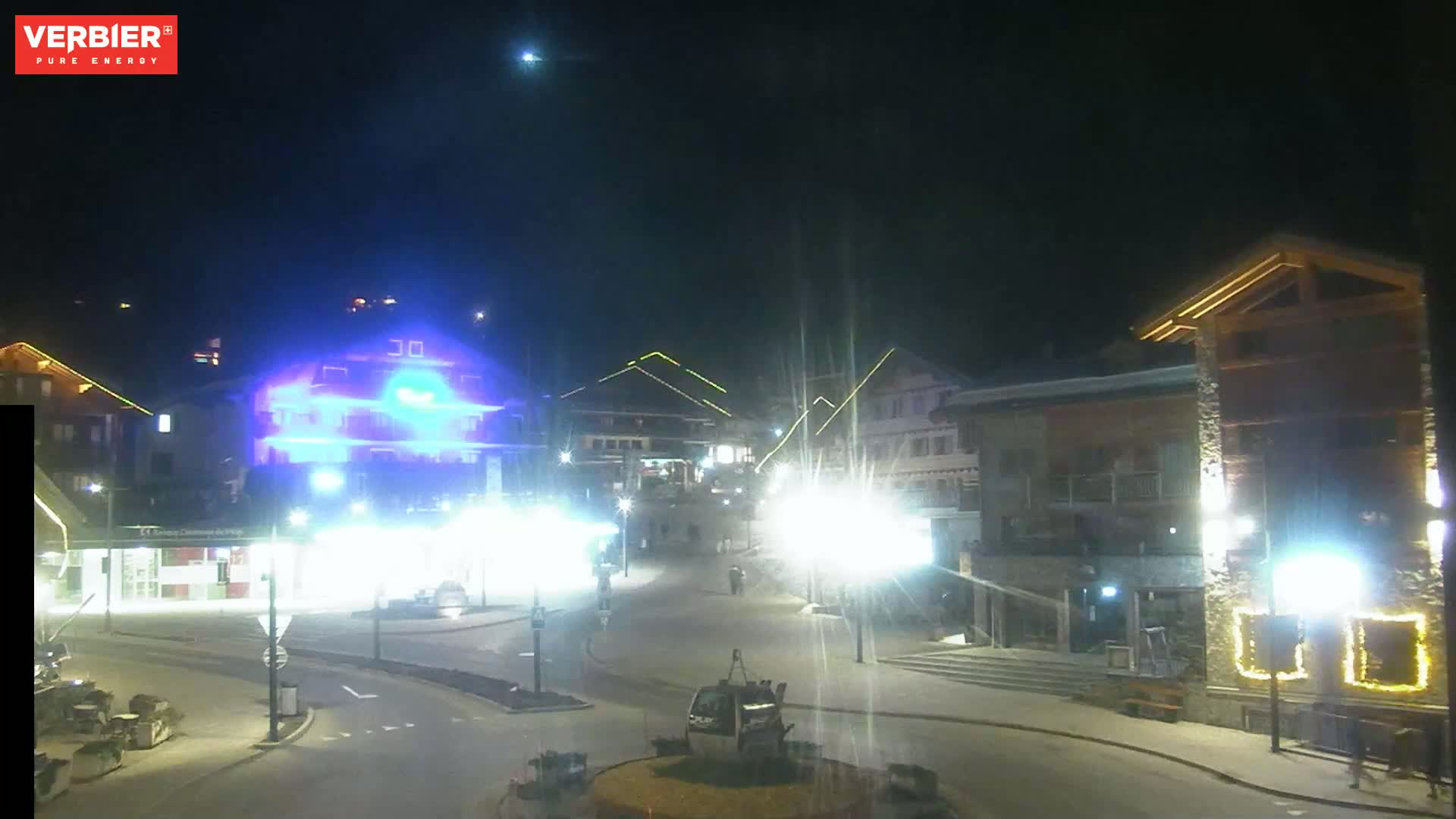 A nighttime view of a town square with brightly lit buildings and a gondola lift, under a clear sky.