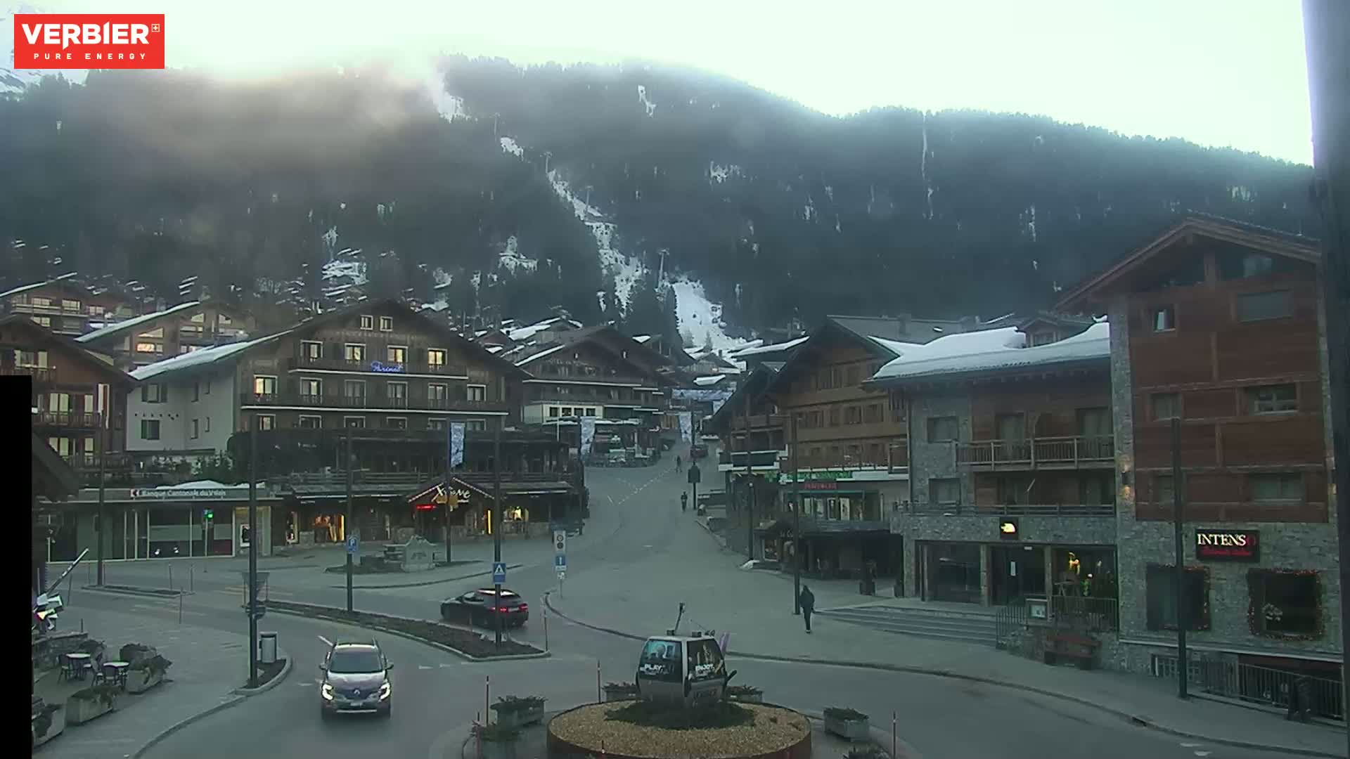 A mostly empty town square in a snow-dusted mountain village on an overcast day, with several multi-story buildings and a few cars visible.