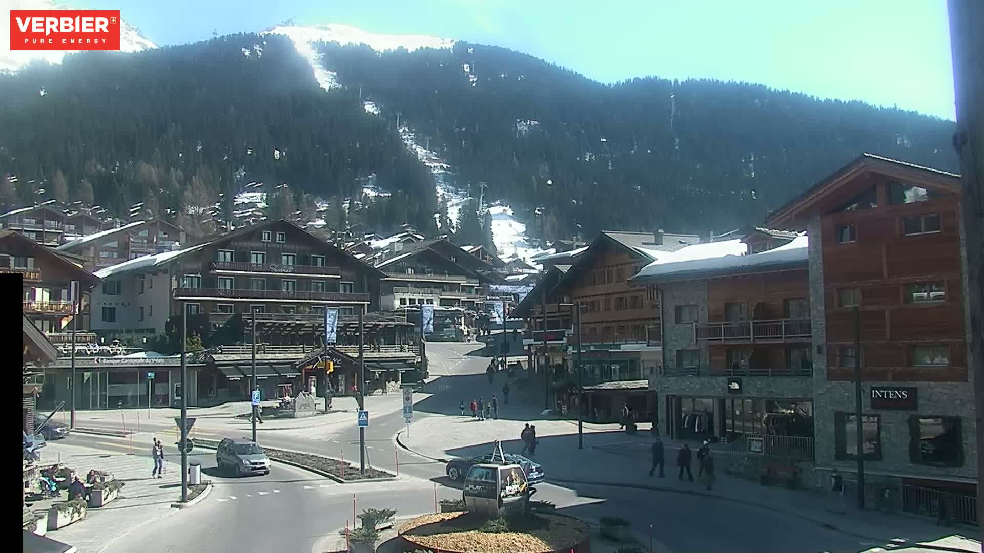 A sunny day in a mountain village, with snow-capped hills in the background and several people walking around a town square.