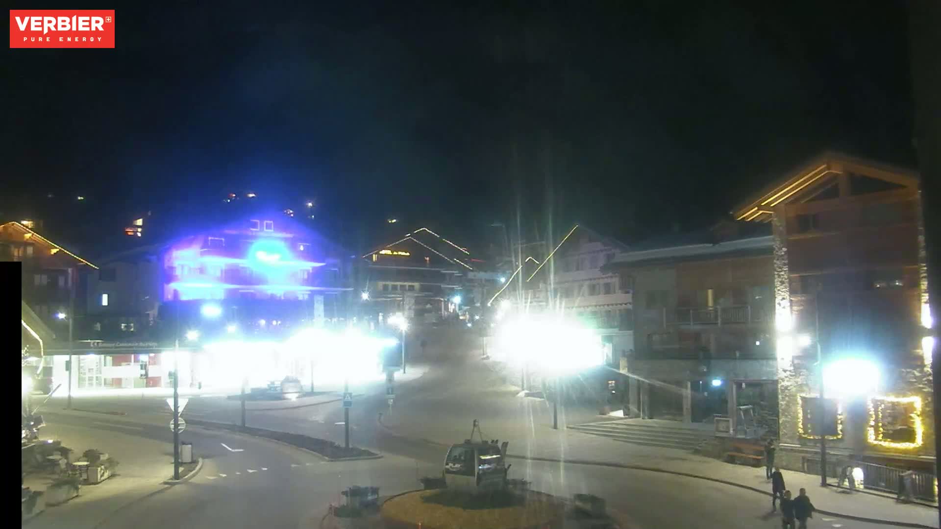 A brightly lit town square at night, with several buildings and a gondola lift, under a clear, dark sky.