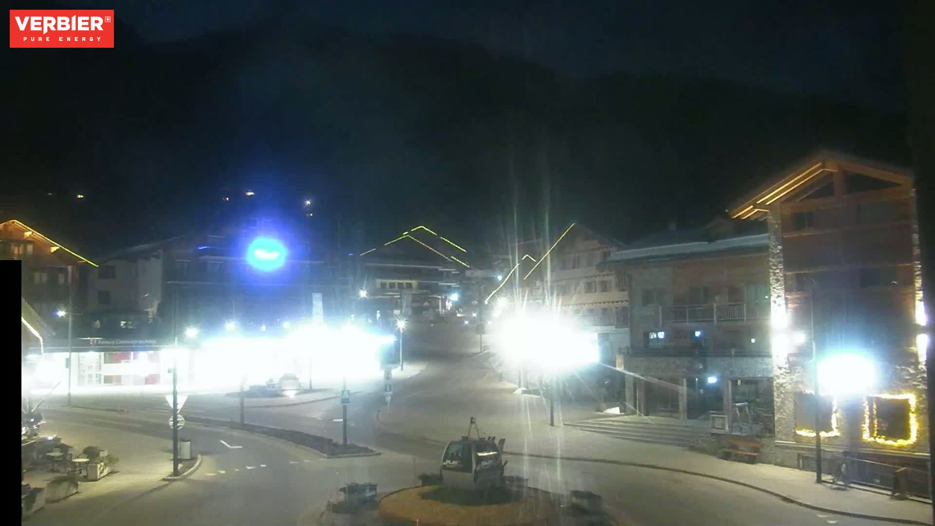 A nighttime view of a snow-covered town square with brightly lit buildings and a gondola lift in the center, under a dark sky.