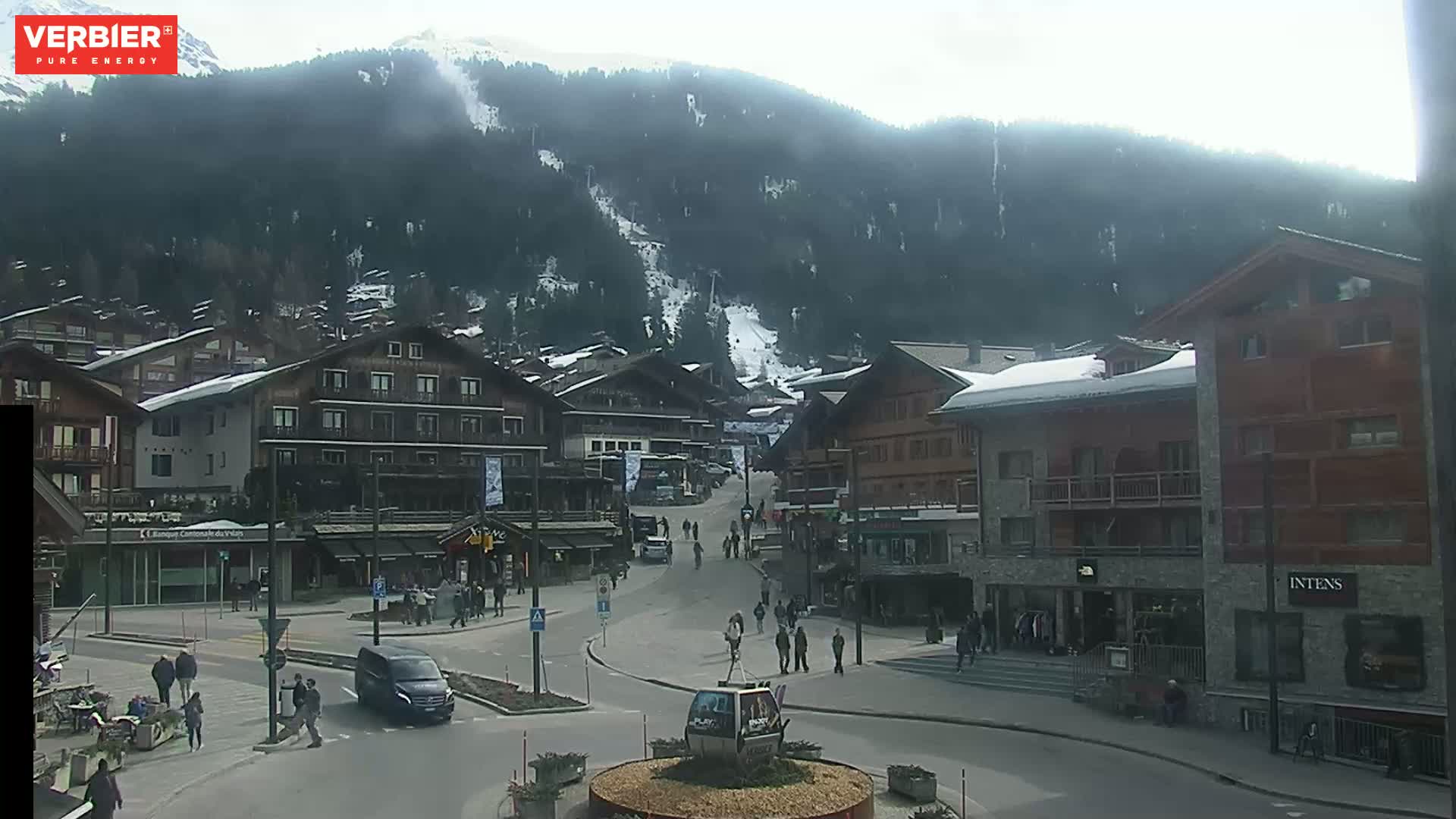 A mostly sunny day in a snow-covered mountain village with several multi-story buildings and people walking around a town square, a ski slope visible on the mountain in the background.