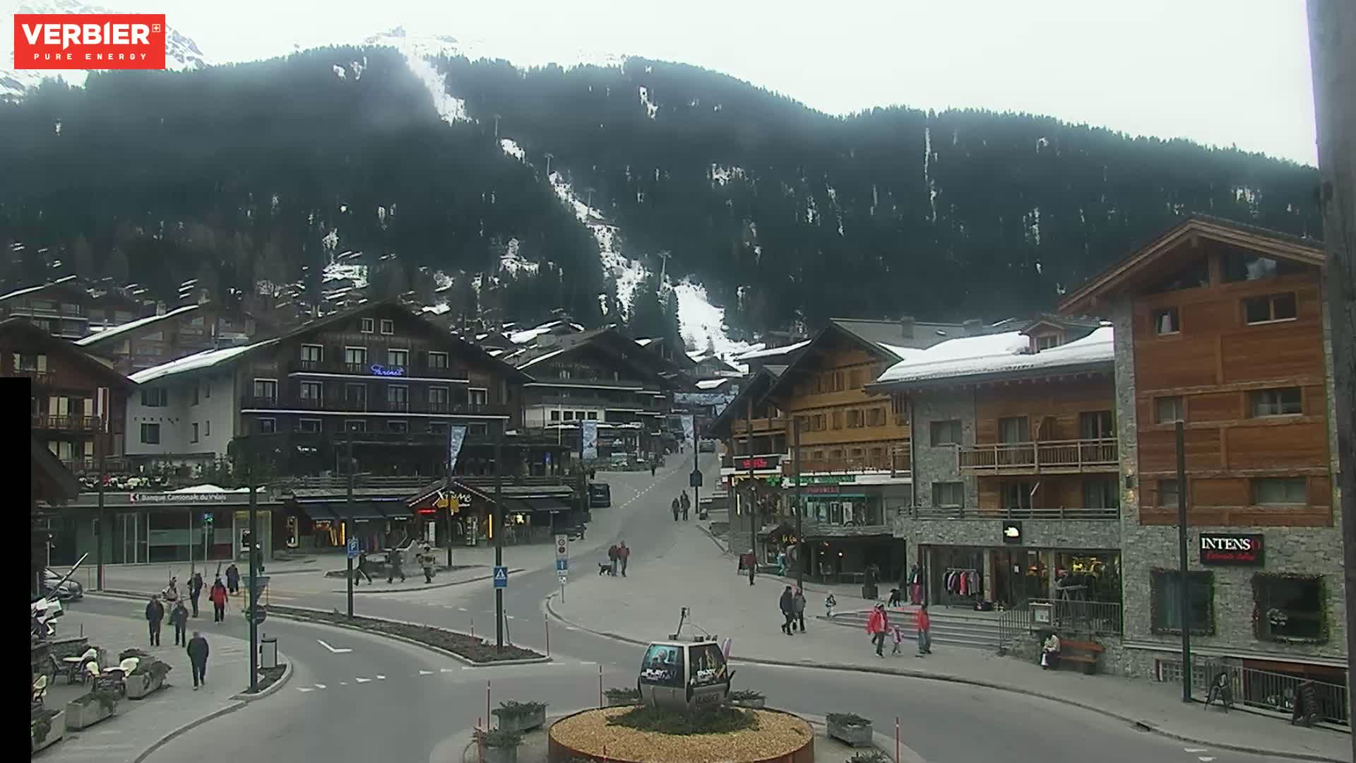 A mostly cloudy day in a snow-covered mountain village with several people walking around a town square, and buildings in chalet style architecture.