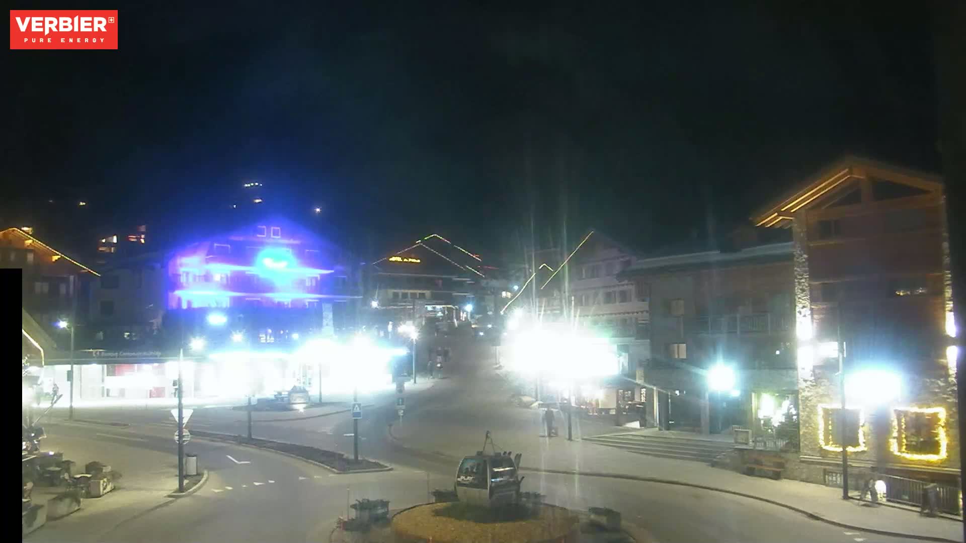 A brightly lit town square at night, with several buildings and a gondola lift visible.