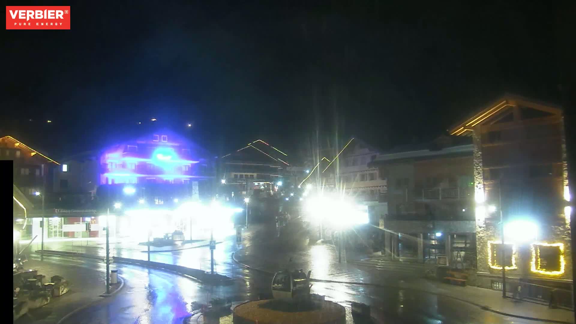 A wet, nighttime scene shows brightly lit buildings and streets in a town square, with some lights reflecting off wet pavement.