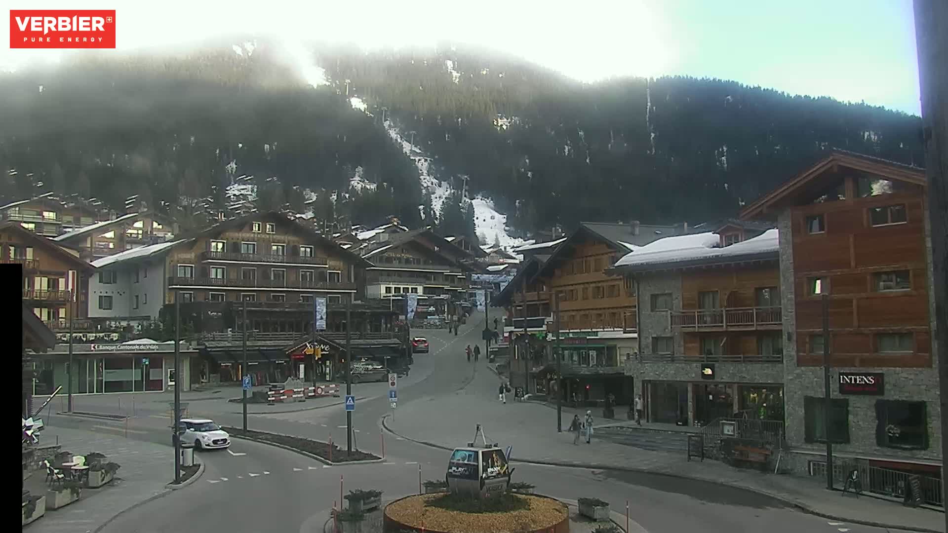 A mountain village square with several buildings, a few cars and pedestrians, and a ski slope visible on the mountainside in the background under hazy conditions.