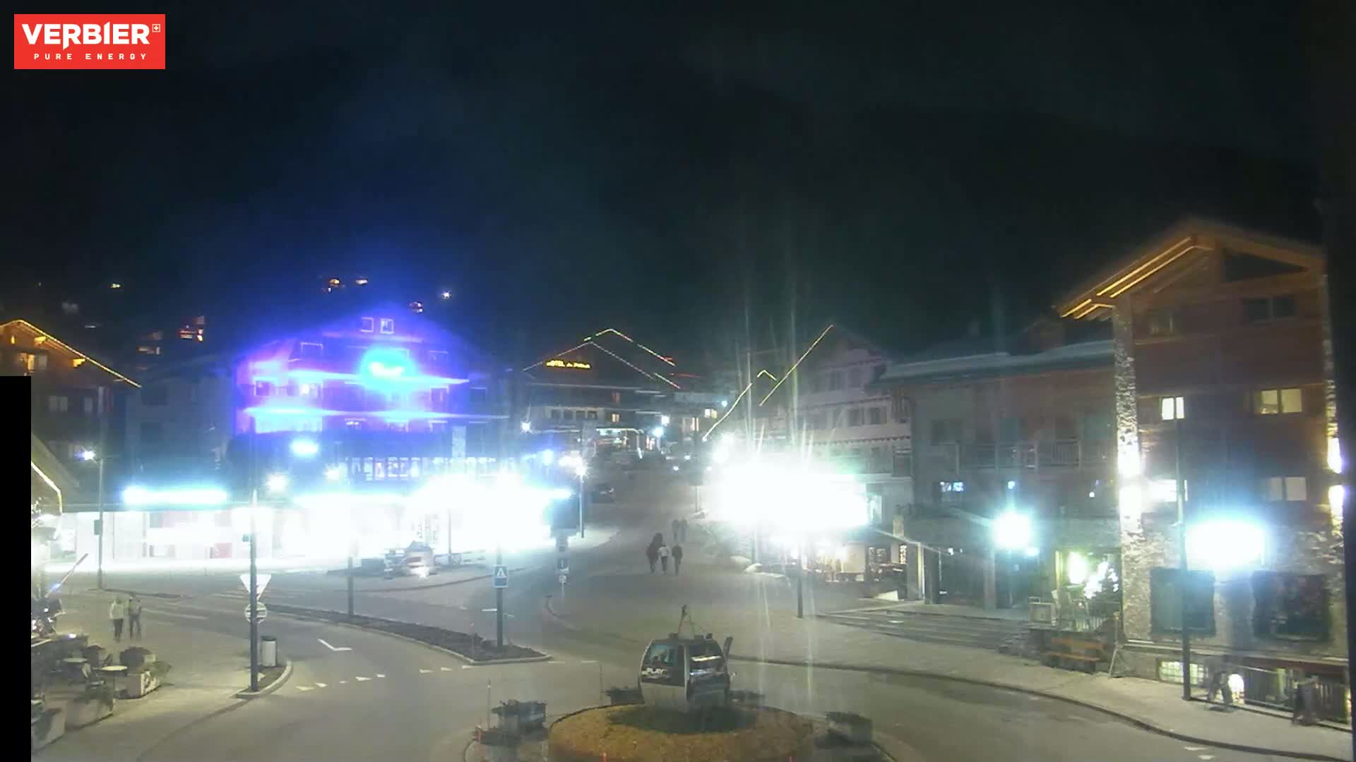 A brightly lit town square at night, with several buildings illuminated and a few people walking around.