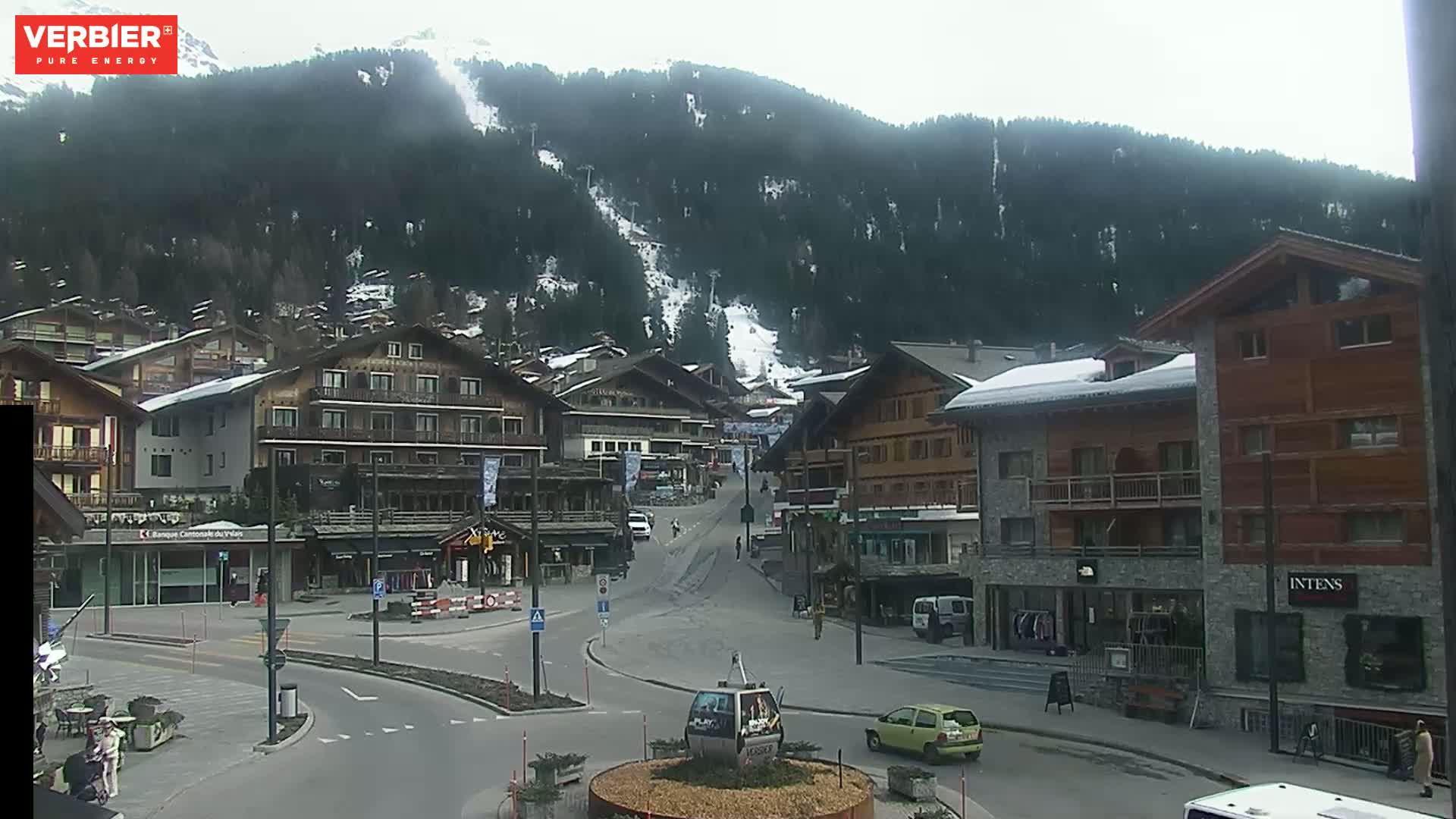 A mostly cloudy day reveals a village nestled at the base of a snow-dusted mountain, with several buildings and a few cars visible in the town square.