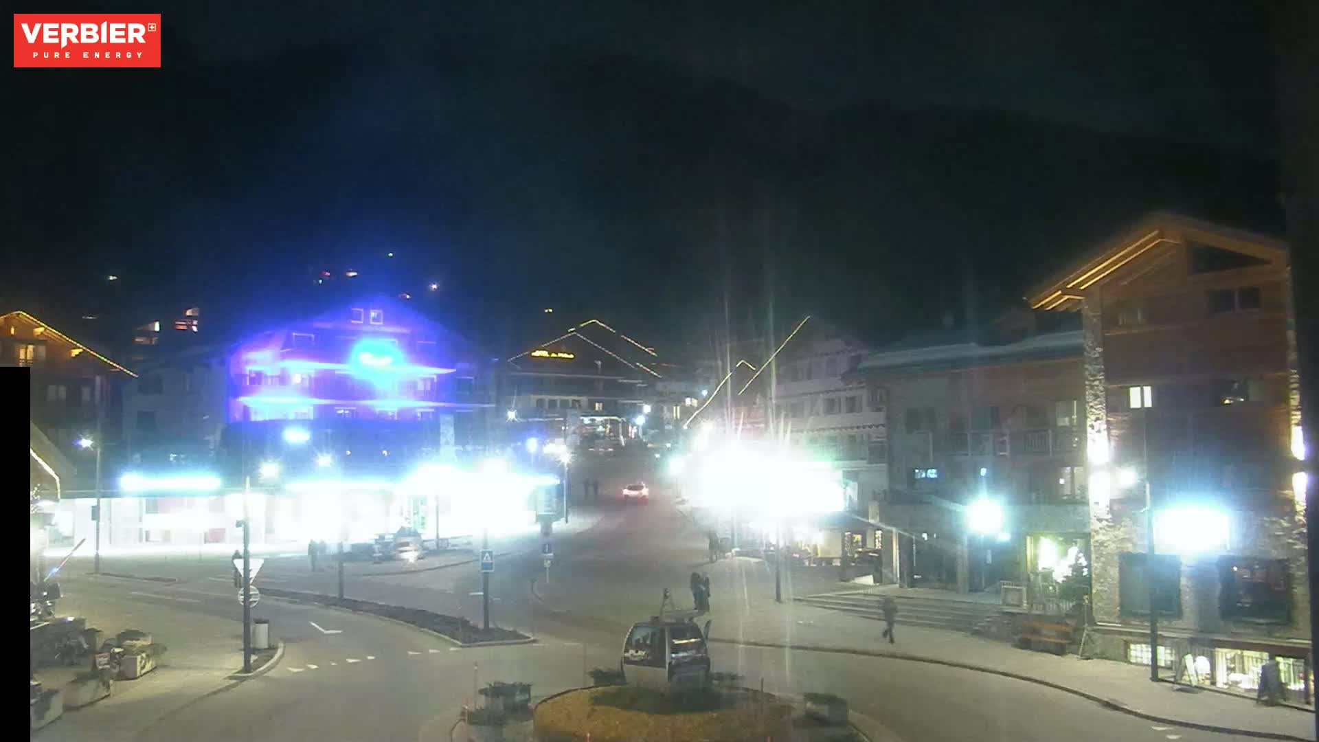 A nighttime, brightly lit town square with several buildings, a few cars, and some pedestrians under a dark sky.
