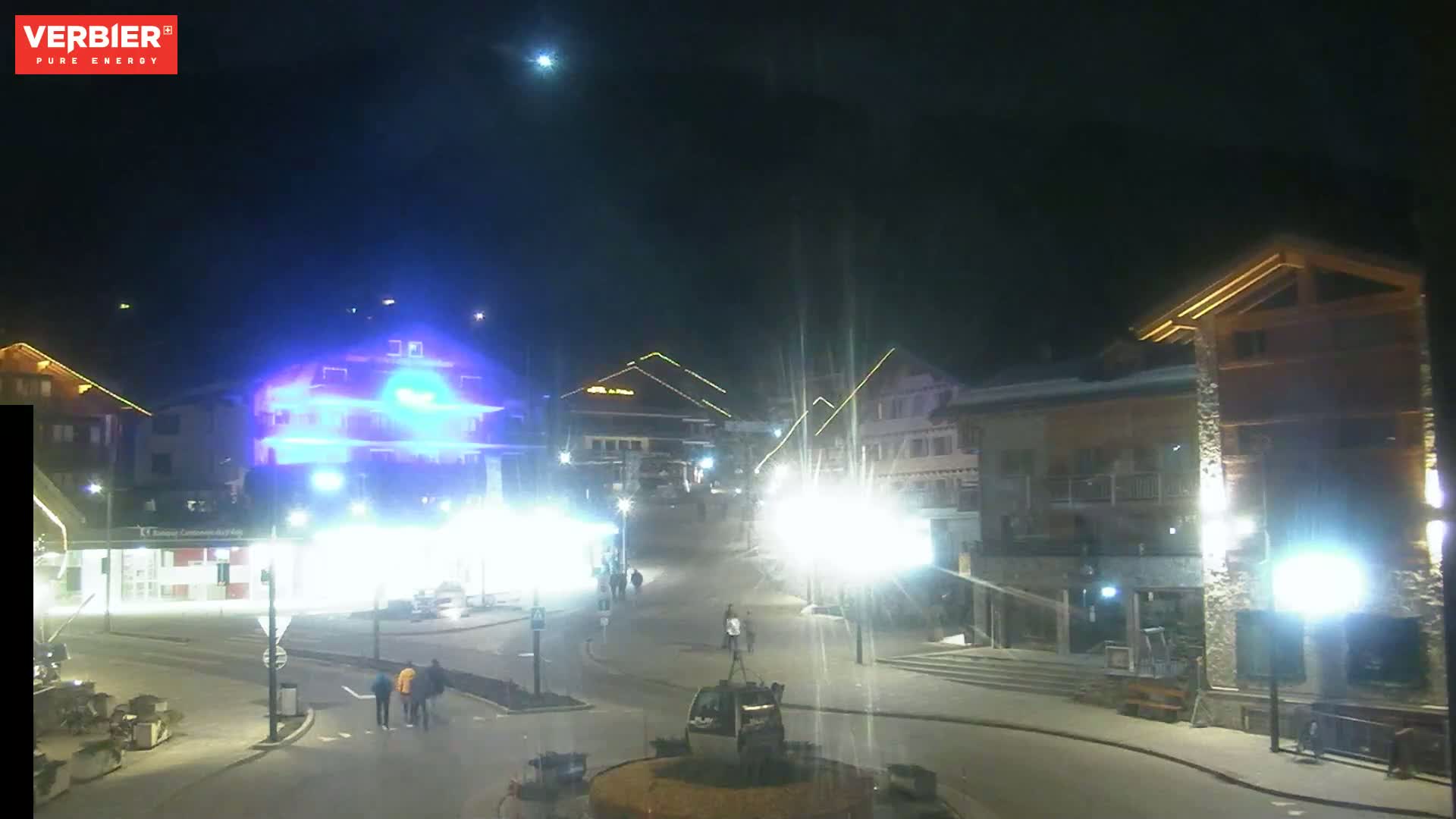 A nighttime, brightly lit town square with several buildings and a few people walking around under a clear sky.