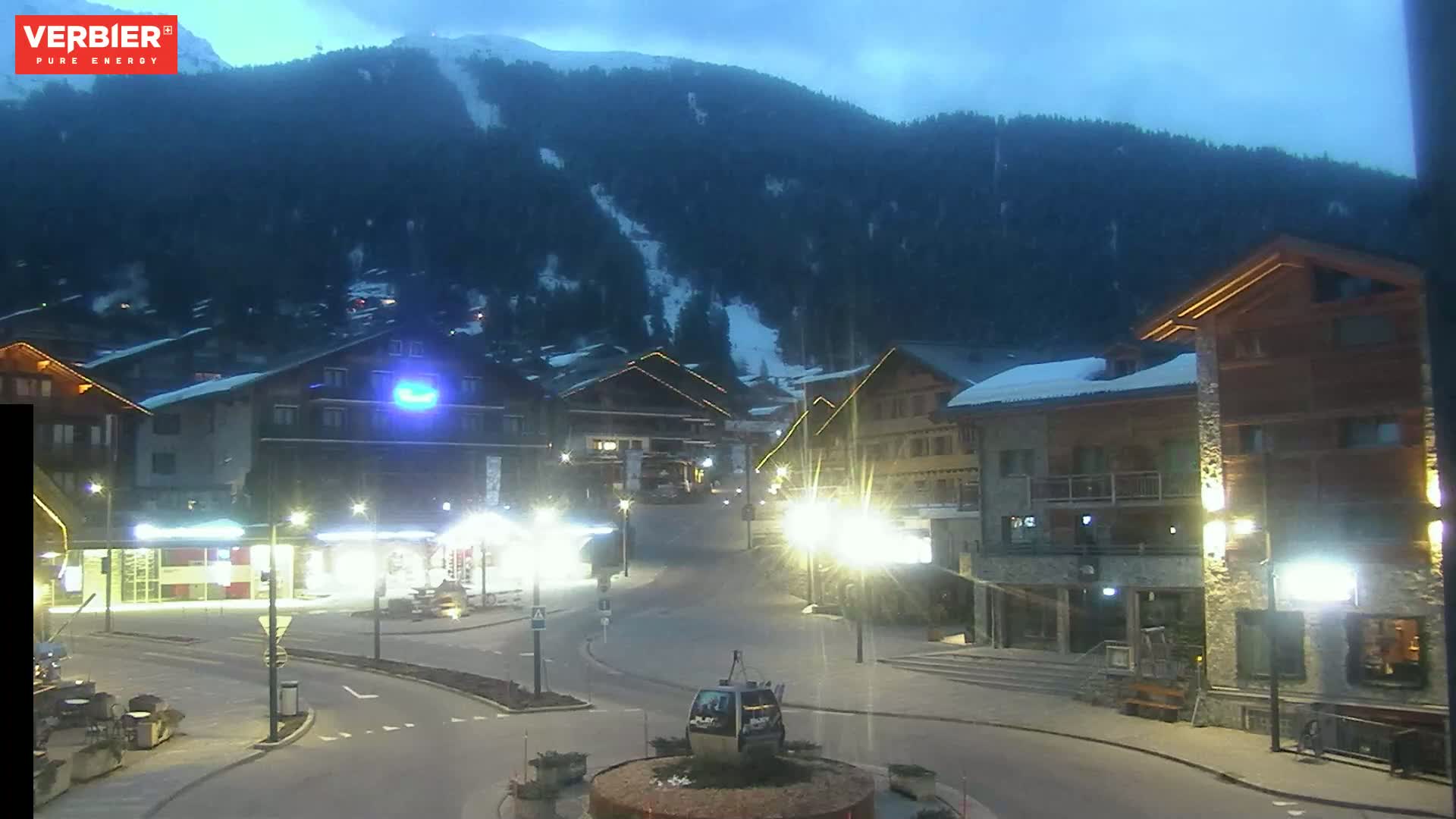 A nighttime, snow-dusted mountain village scene under a twilight sky, showing a mostly empty town square with illuminated buildings and streetlights.