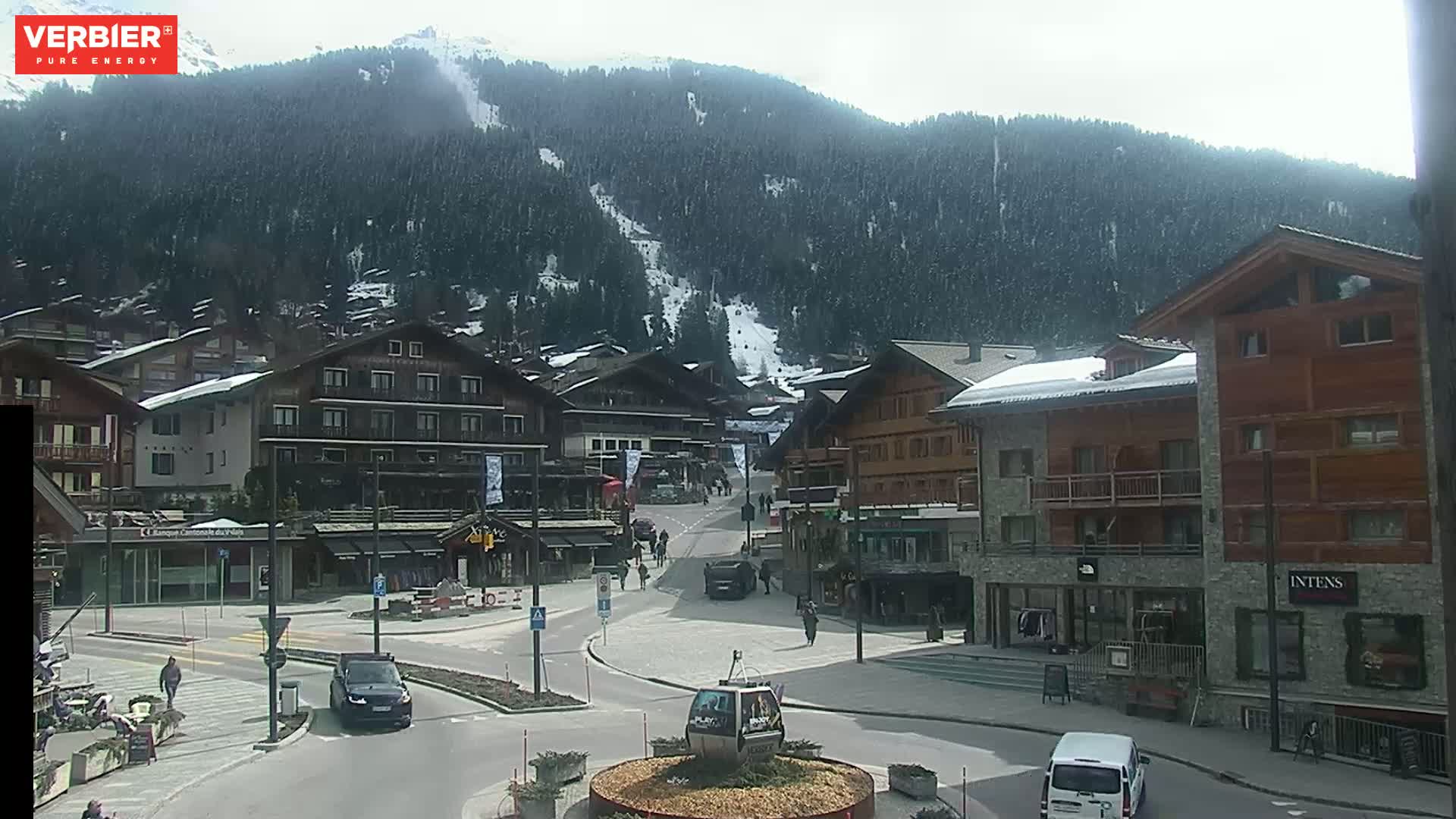 A mostly sunny day reveals a village nestled in a valley, with snow-capped mountains in the background and several cars parked on the streets.