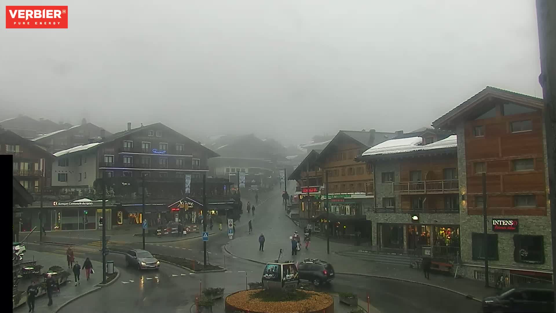 A foggy, rainy day in a village with multi-story buildings and several people walking on the street.