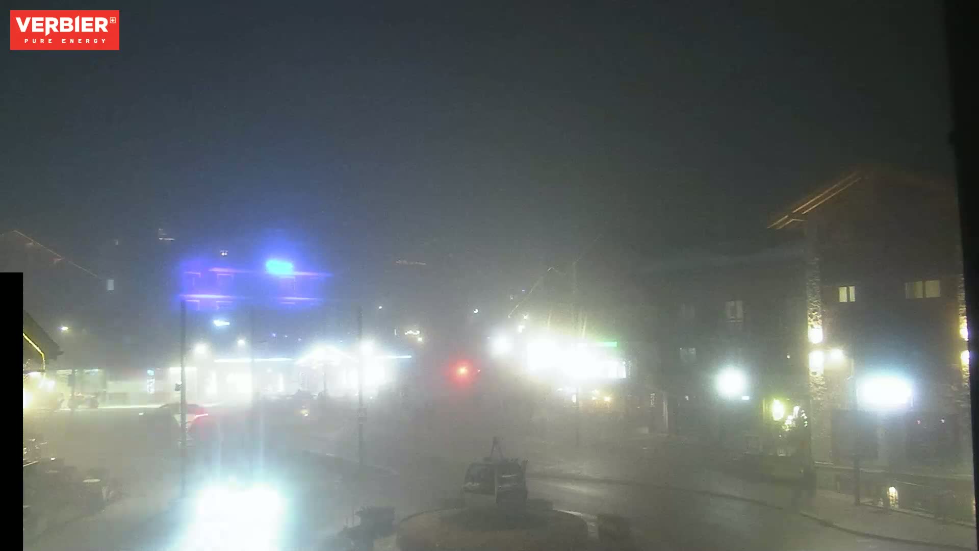 A foggy night scene shows a street with buildings and vehicles, illuminated by streetlights.