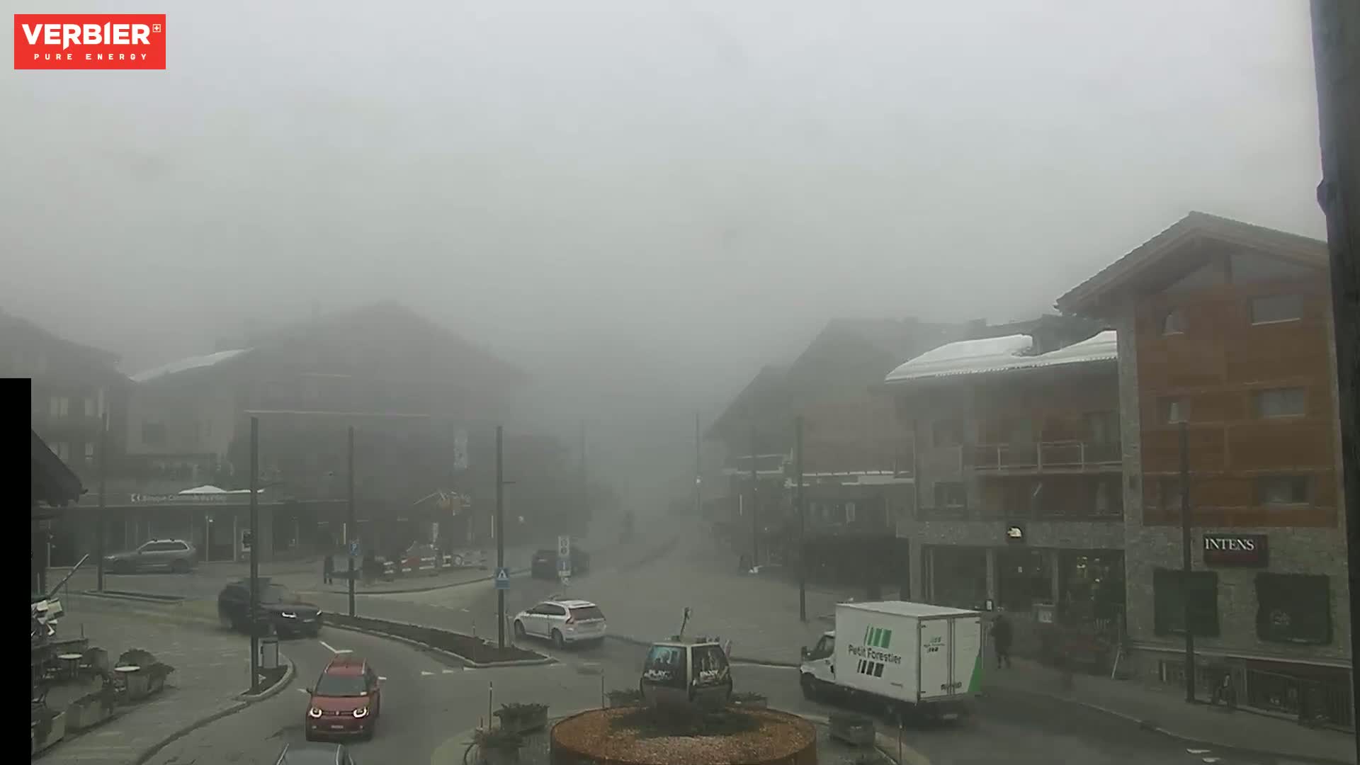A foggy town square with several buildings and a few cars is visible, some with snow on their roofs.