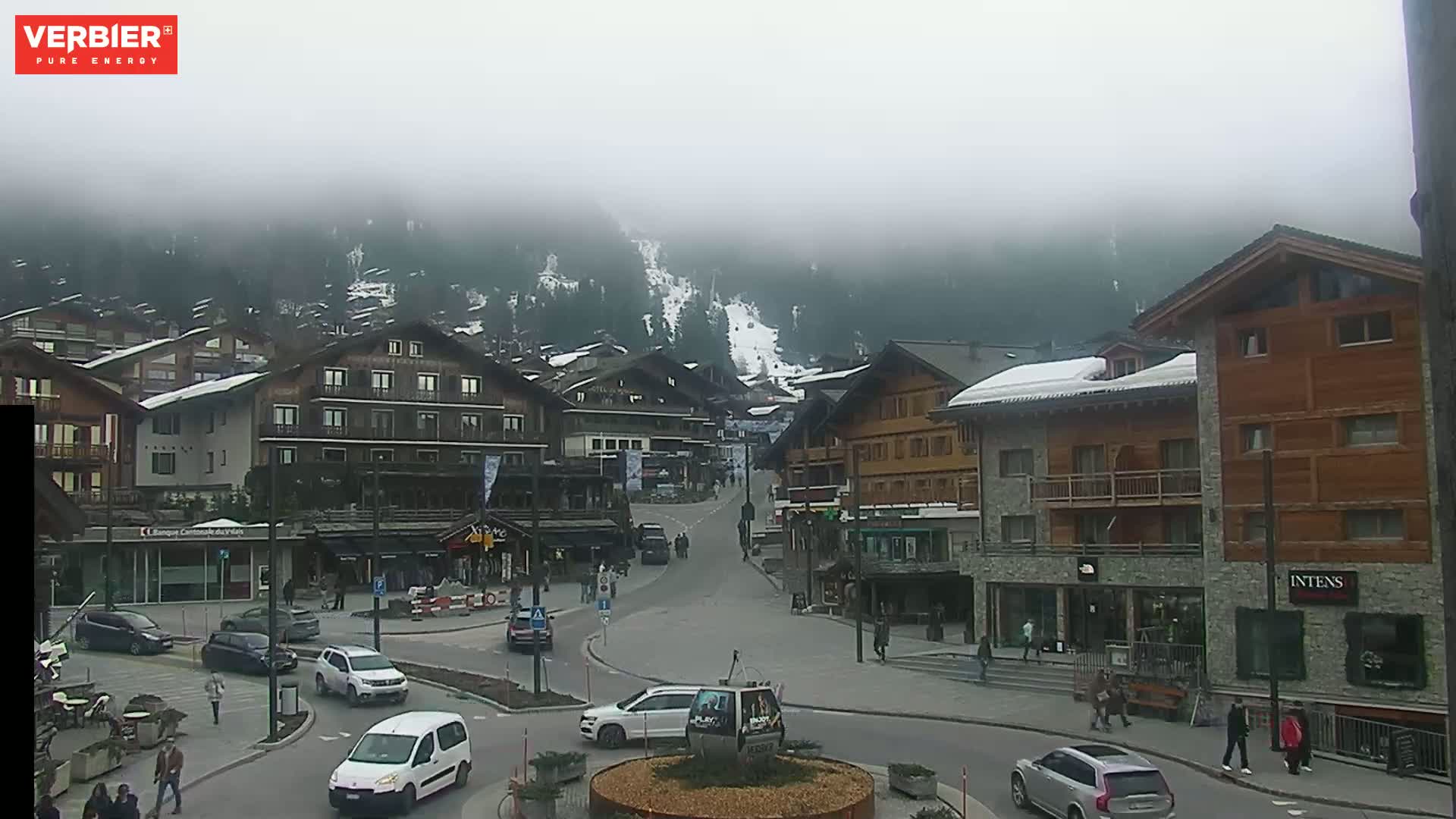 A foggy mountain village street scene with several multi-story buildings and cars, showing a snowy mountainside in the background.