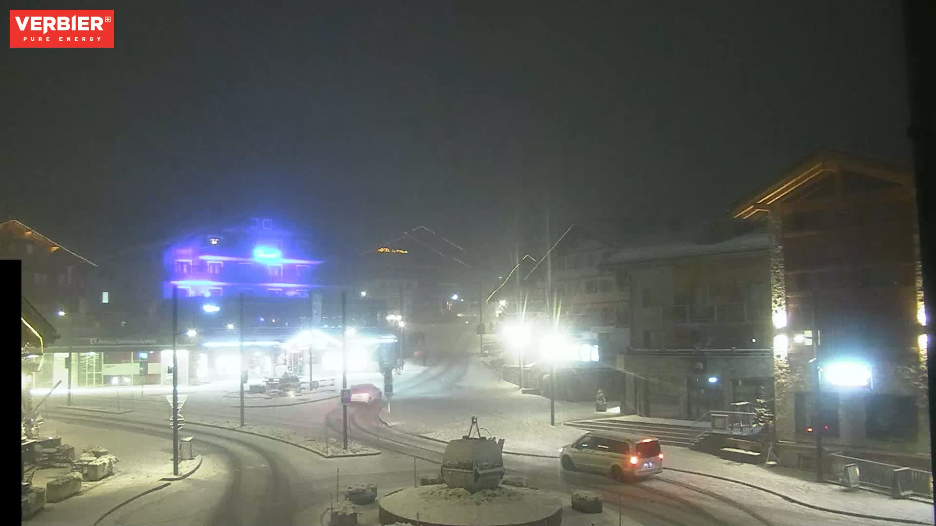 A snow-covered town square at night, illuminated by streetlights, with cars and buildings visible in the falling snow.