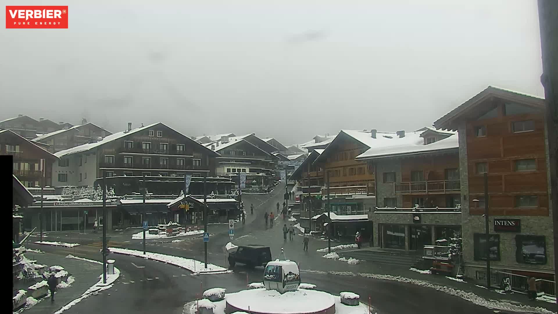 A snow-covered village square with several multi-story buildings, a few pedestrians, and vehicles under an overcast sky.