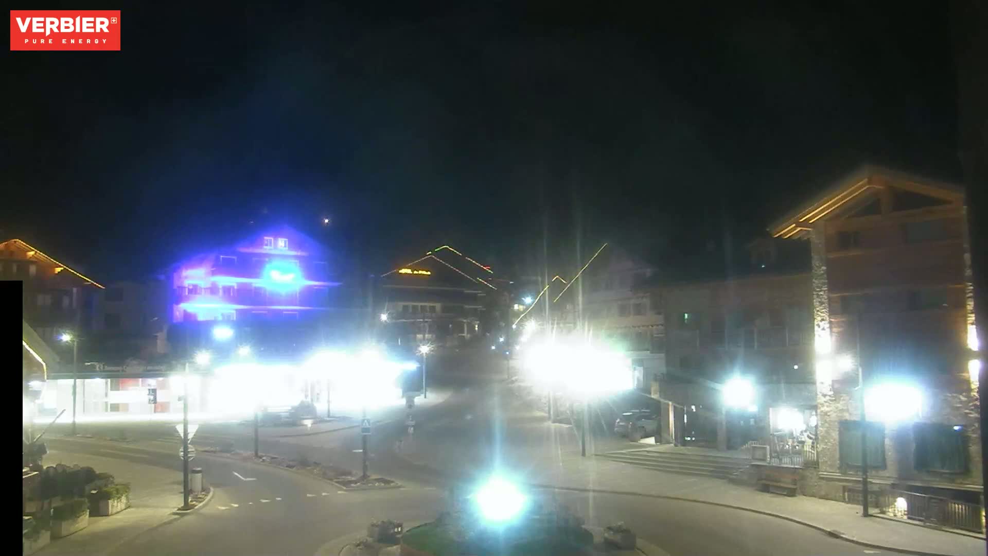 A nighttime, brightly lit town square with several buildings and streetlights.