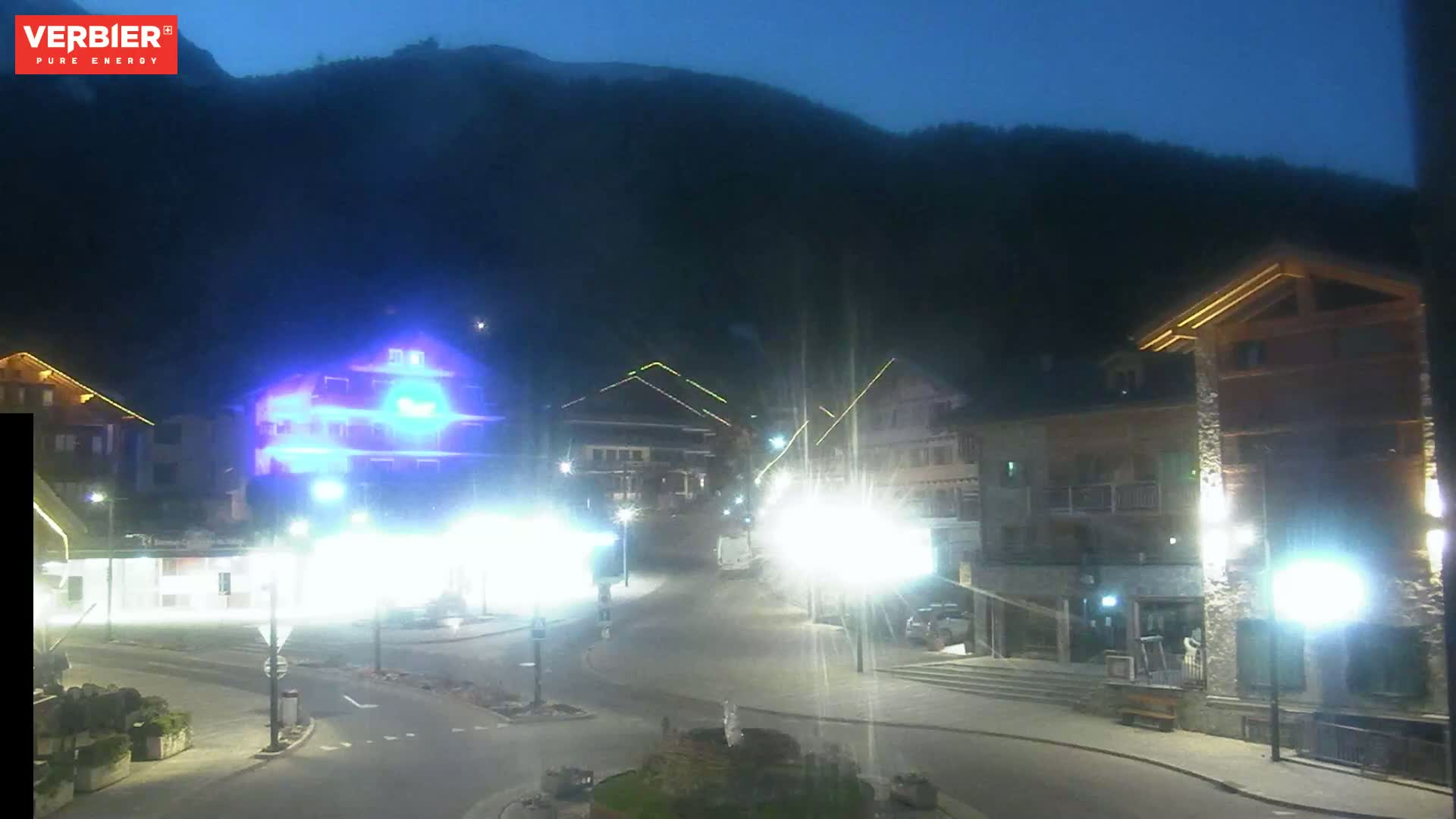 A nighttime view of a village square nestled in a mountain valley under a dark, clear sky, illuminated by streetlights.