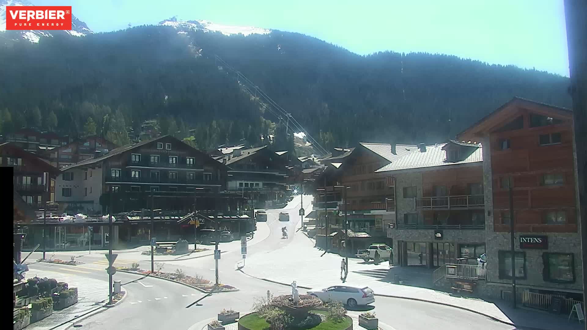 A sunny mountain town square with several buildings, a few cars, and people walking, surrounded by a mountainous landscape with some snow on the peaks.