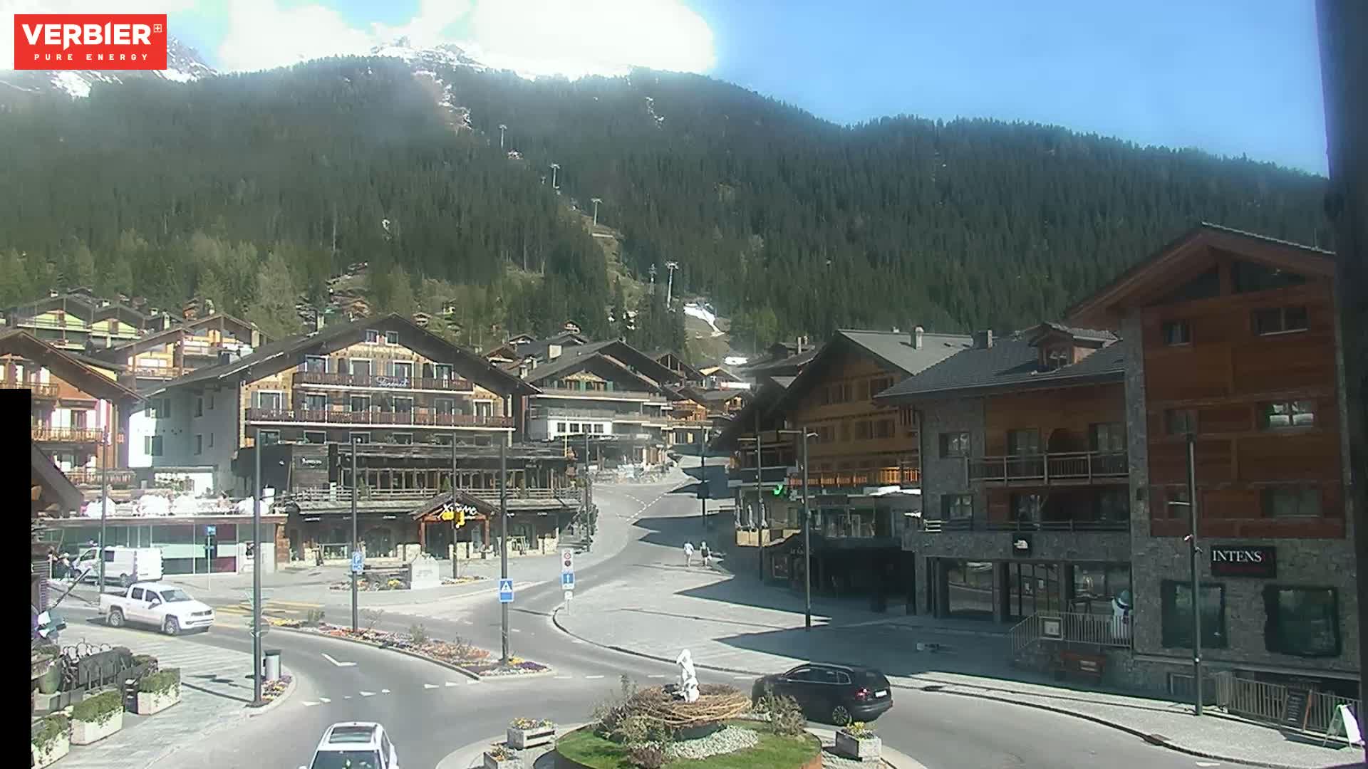 A sunny mountain village square with several multi-story buildings and cars, nestled against a backdrop of a forested mountain range.