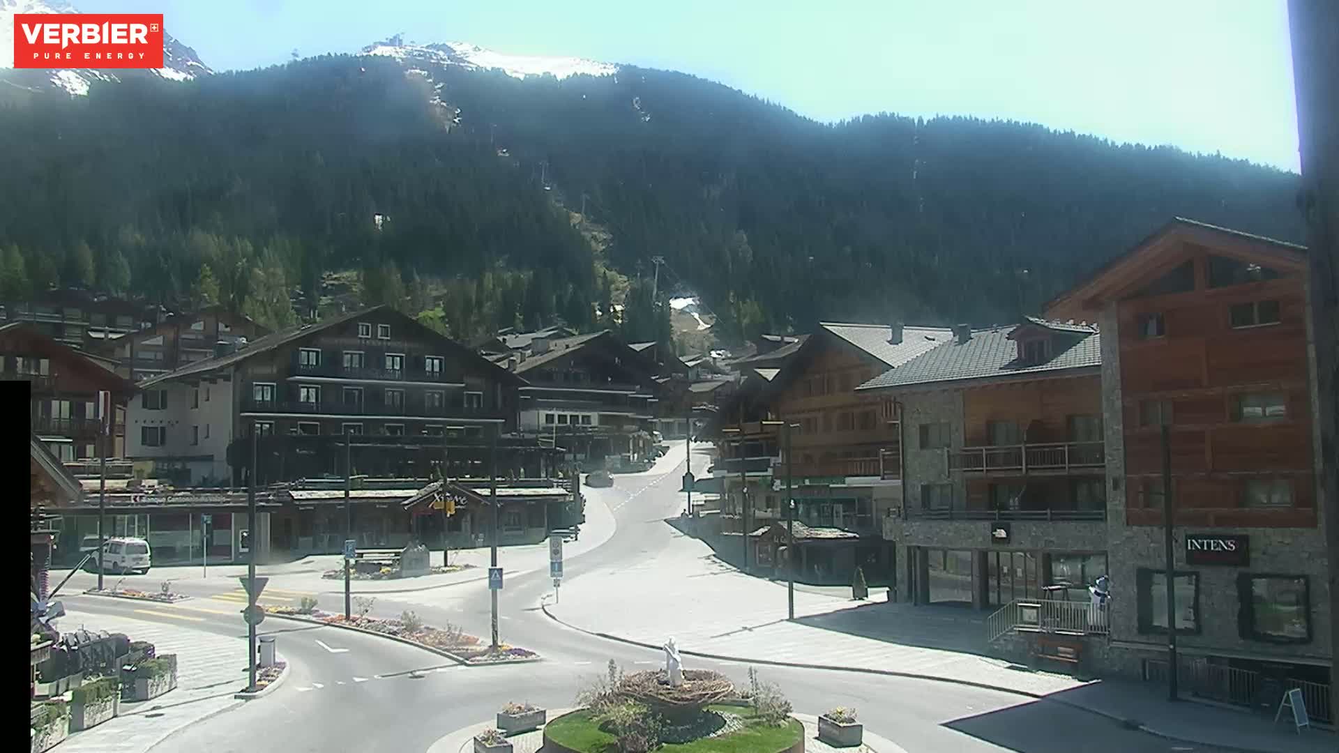 A sunny mountain village square with several buildings, a road curving through it, and a mountain range in the background.